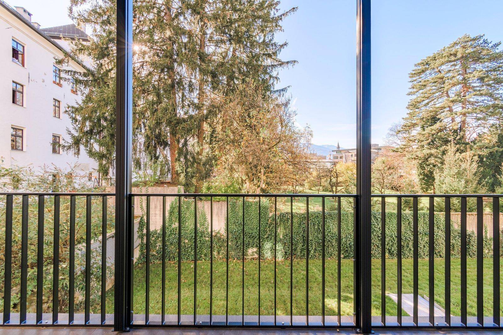 Blick vom Balkon auf einen gepflegten Garten mit hohen Hecken und Bäumen sowie angrenzenden Gebäuden.