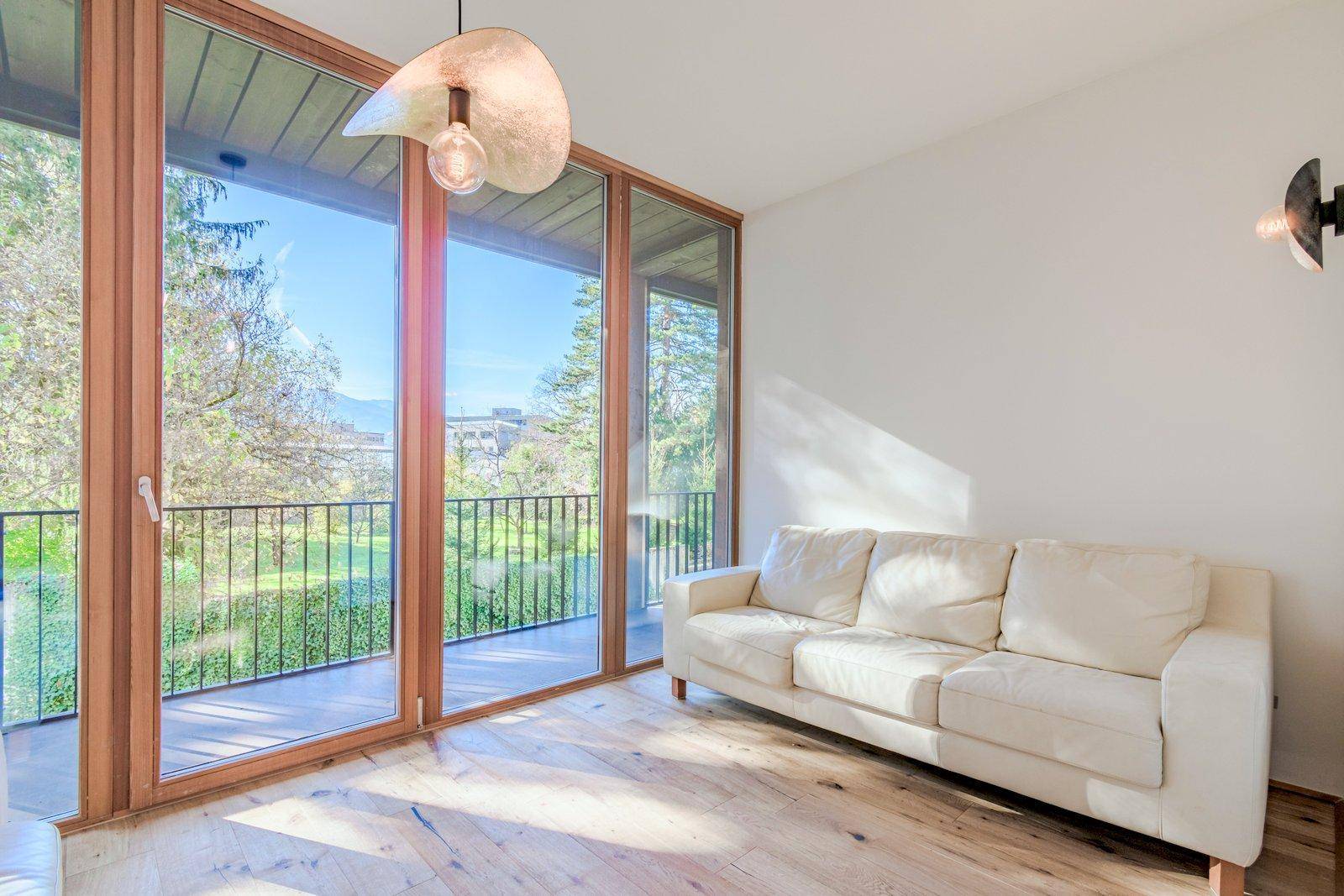 Helles Wohnzimmer mit weißem Sofa, großen Fenstern und Blick ins Grüne sowie Zugang zum Balkon.