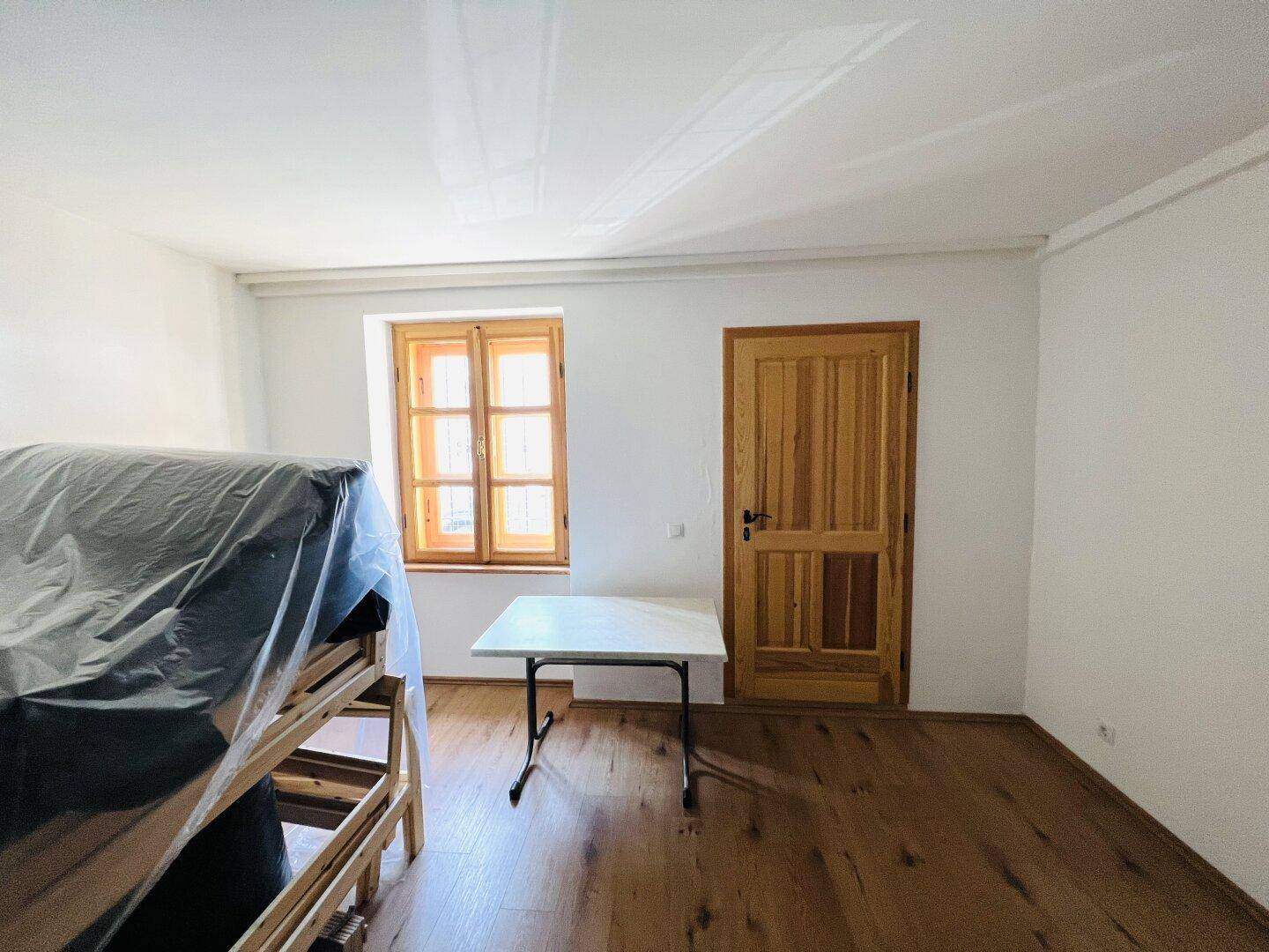 Helles Zimmer mit Holzboden, einem Fenster und einer Holztür, bereit zur Einrichtung.