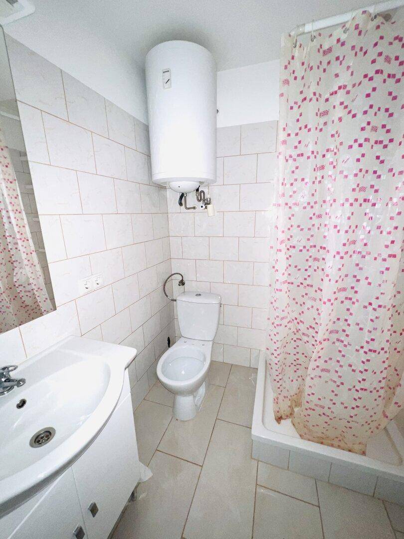 Kleines Badezimmer mit weißen Fliesen, Dusche und Warmwasserboiler an der Wand.