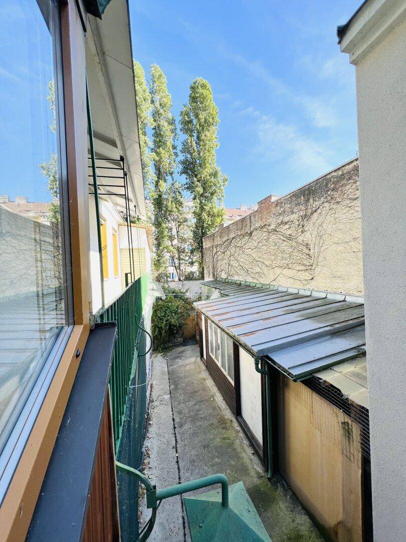 Blick aus der verglasten Loggia auf den schmalen Hinterhof mit hohen Bäumen.