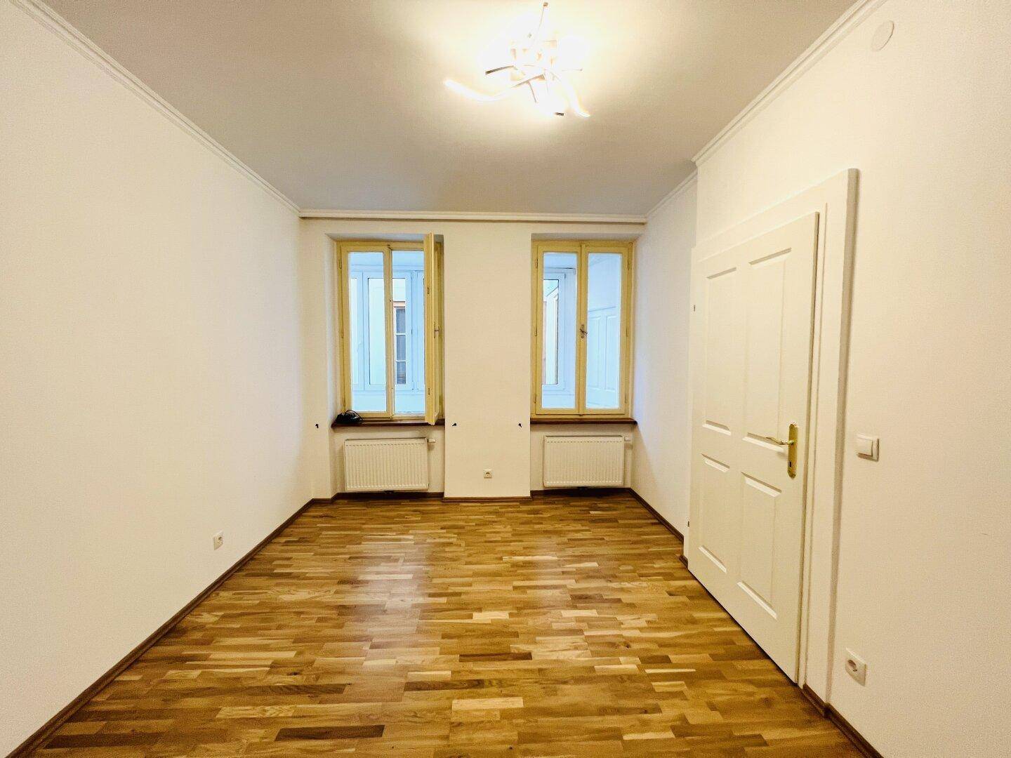 Helles Zimmer mit Parkettboden und zwei Fenstern, bereit zur individuellen Gestaltung.