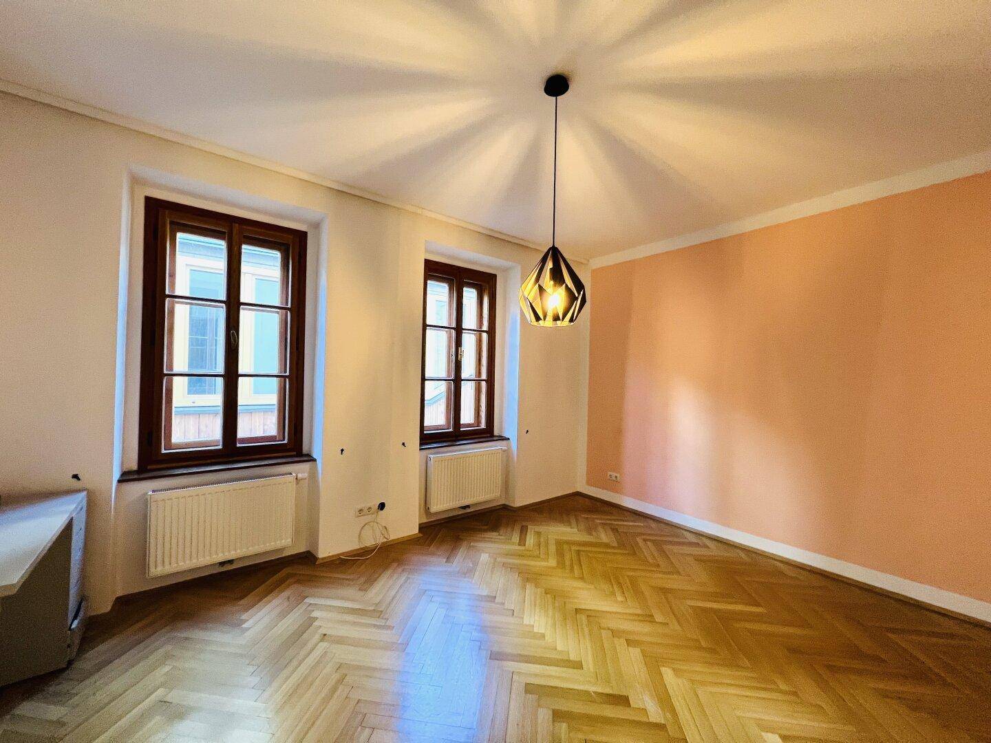 Geräumiges Zimmer mit Fischgrätparkett, zwei Fenstern und einer farbigen Akzentwand.