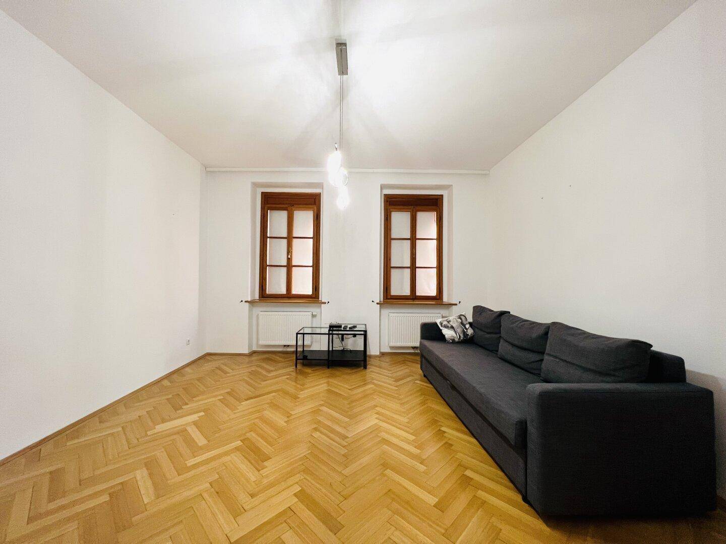 Helles Wohnzimmer mit Fischgrätparkett, bequemer Couch und zwei großen Fenstern.