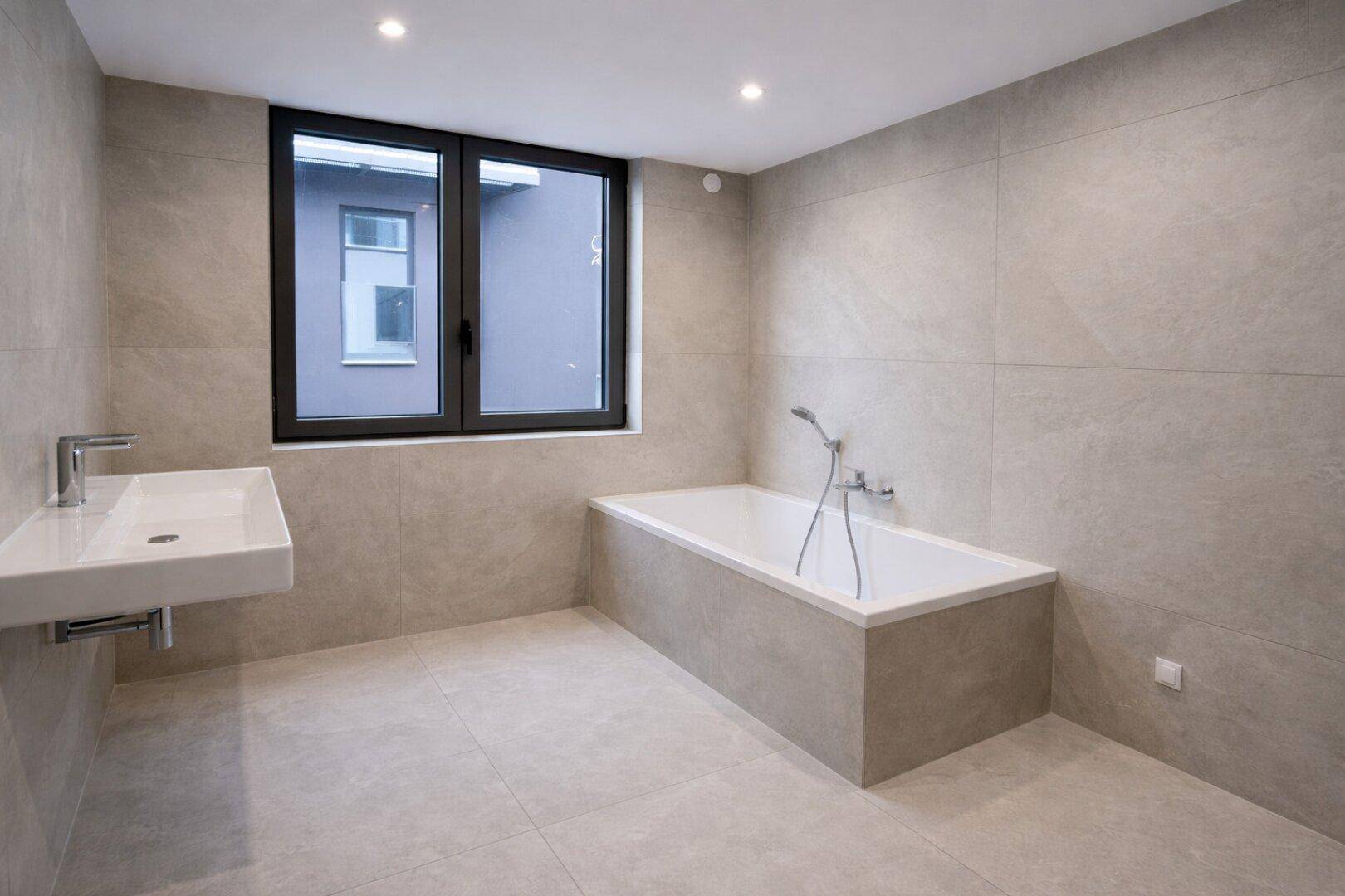 Modernes Badezimmer mit großen Fliesen, Badewanne und einem Fenster für Tageslicht.