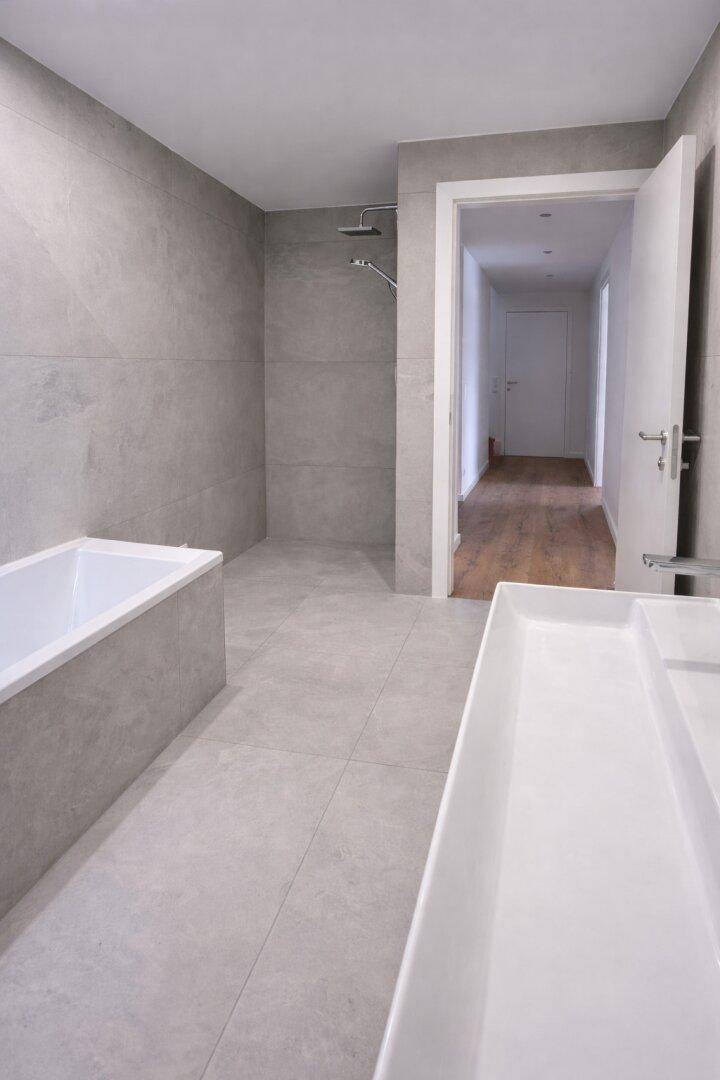 Helles Badezimmer mit modernen grauen Fliesen, Badewanne und einer Duschvorrichtung.