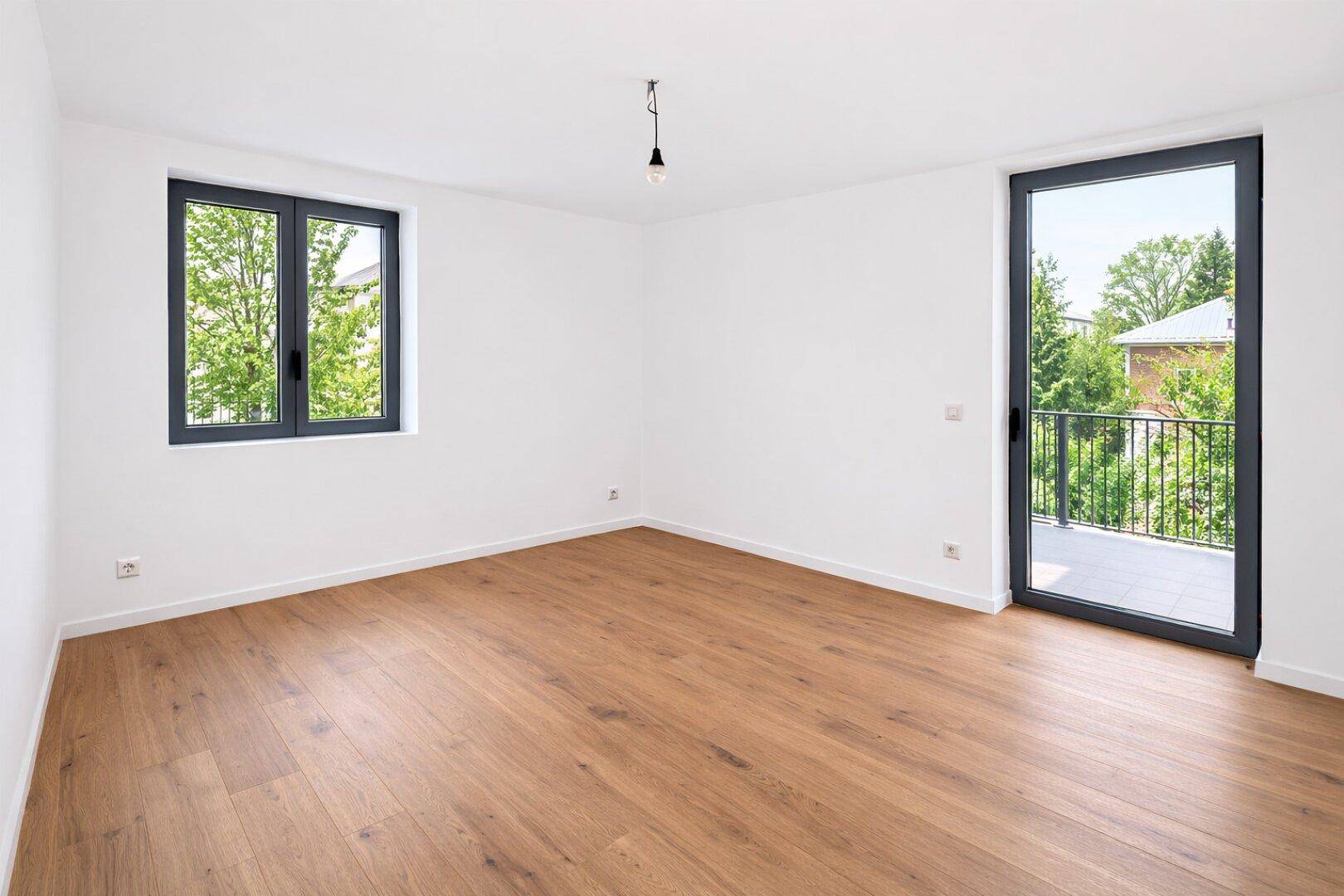 Helles Zimmer mit Holzboden und Zugang zu einem kleinen Balkon mit Blick ins Grüne.