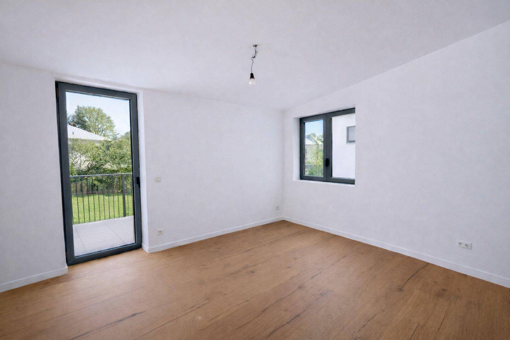 Leeres Zimmer mit Holzboden, großen Fenstern und direktem Zugang zum Außenbereich.