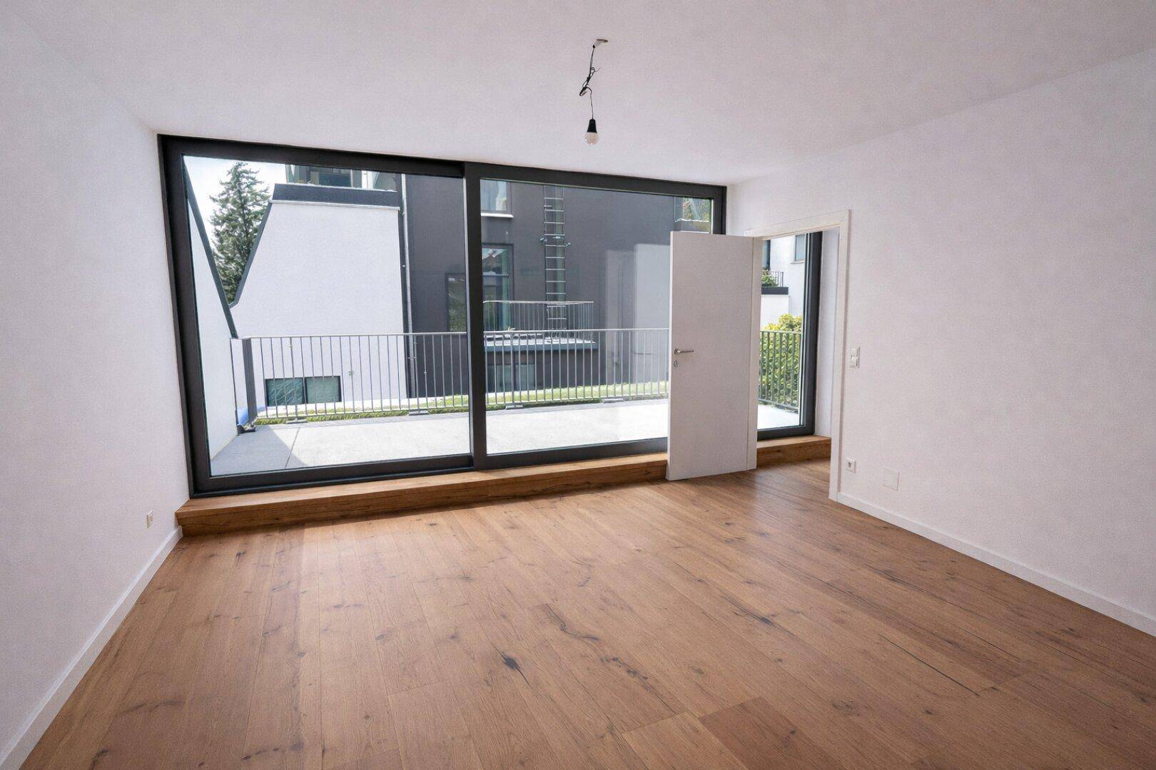 Geräumiger, heller Raum mit Holzboden und breiter Fensterfront zu einer großen Terrasse.