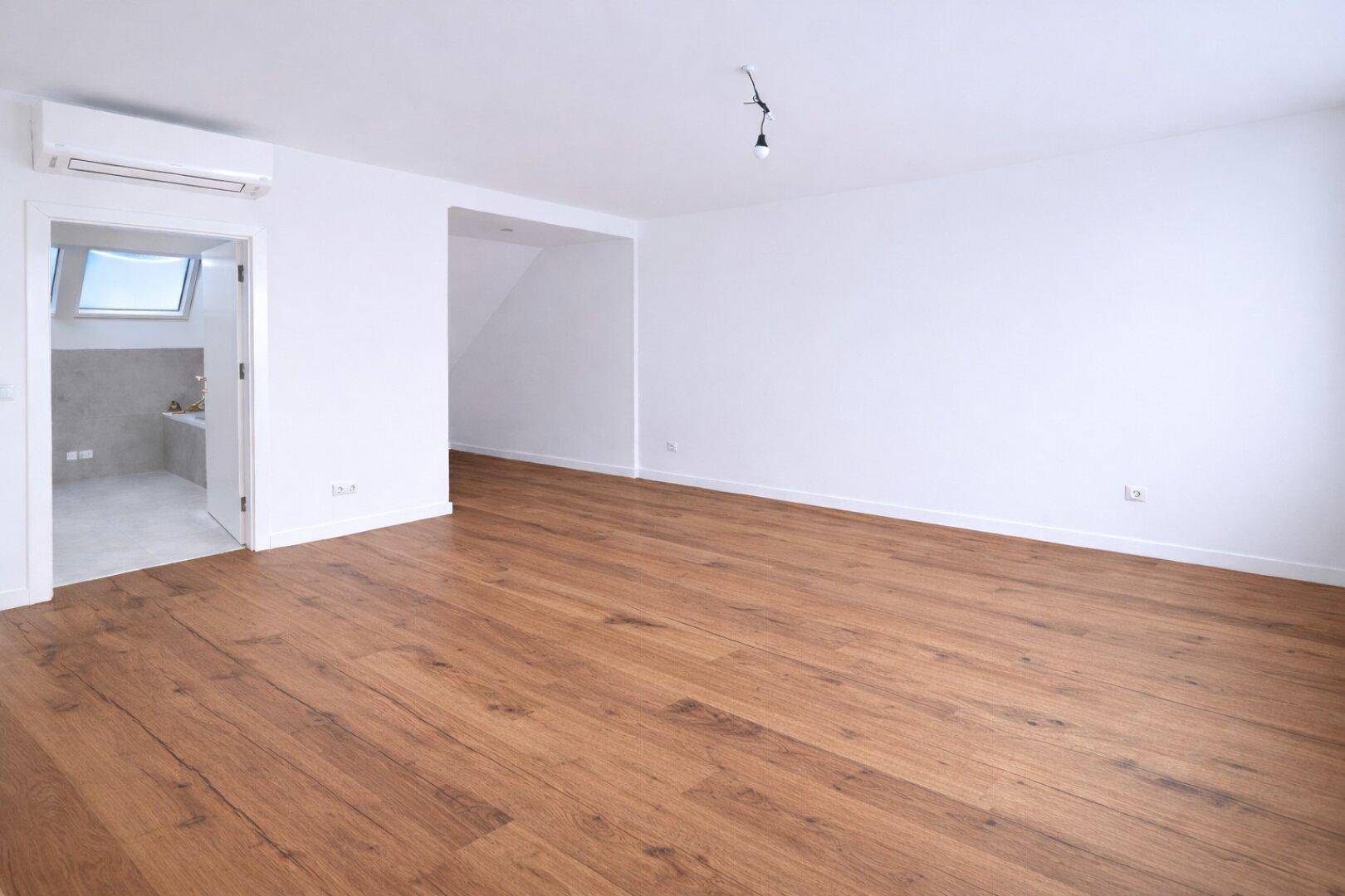 Großer, heller Raum mit Holzboden und Klimaanlage, angrenzend an ein Badezimmer.