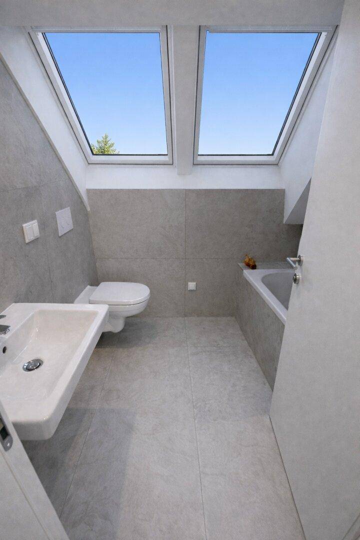 Modernes Badezimmer im Dachgeschoss mit Badewanne, WC, Waschbecken und zwei Dachfenstern.