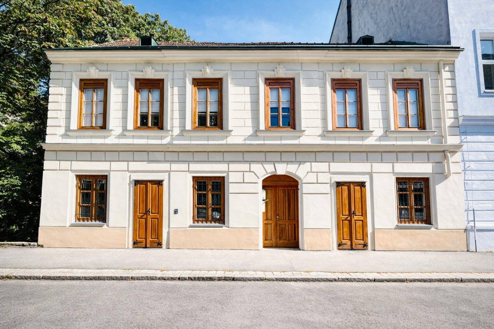 Gut erhaltene Altbaufassade mit traditionellen Holzfenstern und Holztüren, umgeben von Grün.