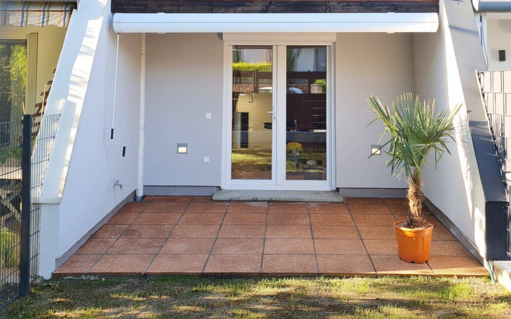Sonniger Terrassenbereich mit Fliesenboden und Zugang zum Innenbereich, umgeben von einer kleinen Grünfläche.