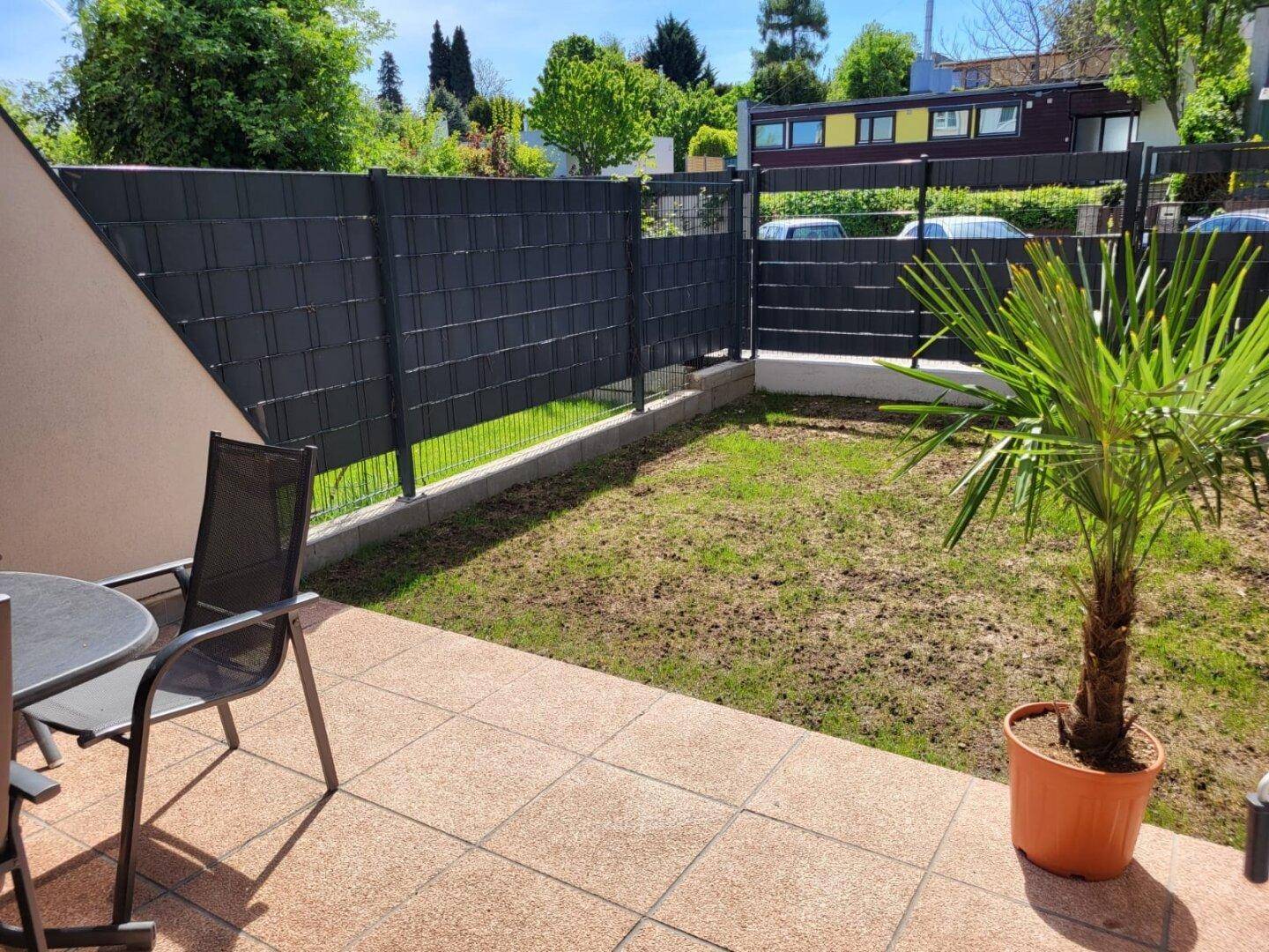 Sonniger Terrassenbereich mit Fliesenboden und Sitzgelegenheiten, umgeben von einer kleinen Grünfläche.