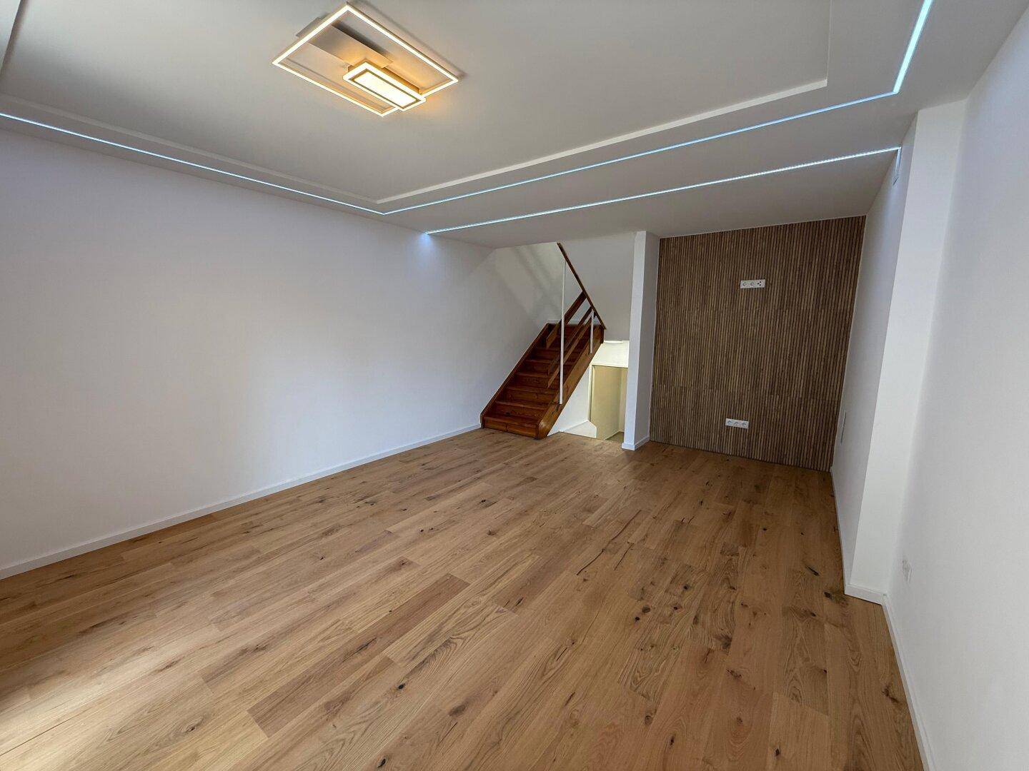 Offener Wohnbereich mit elegantem Holzfußboden und moderner Deckenbeleuchtung, verbunden mit einer Holztreppe.