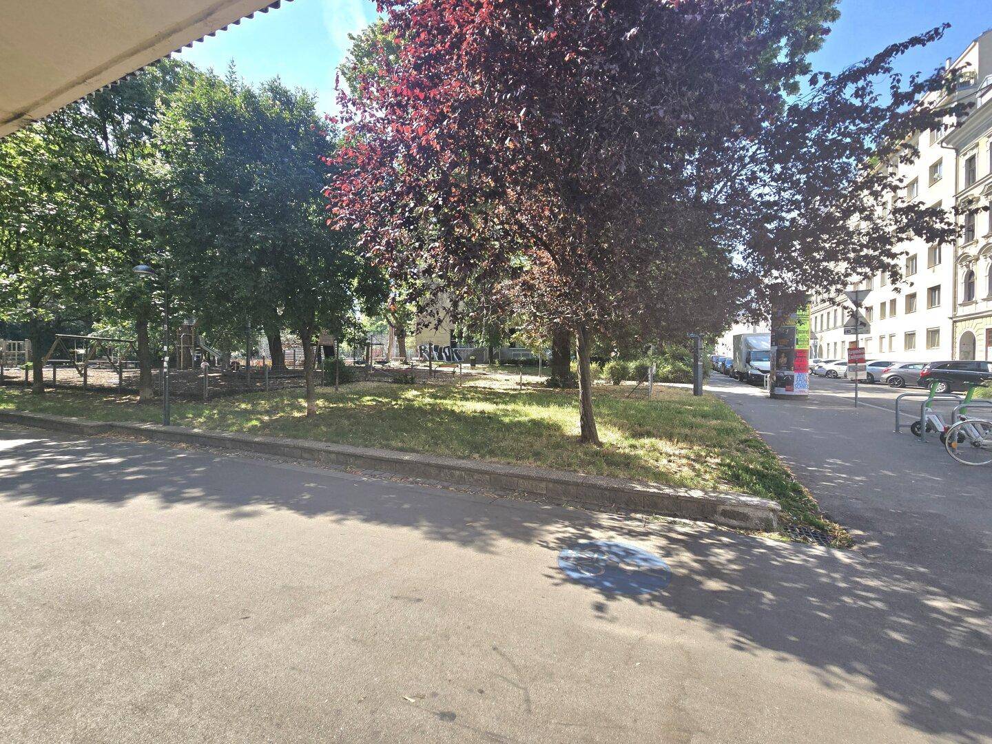 Grünfläche mit Bäumen und Spielgeräten neben einer Straße mit parkenden Autos.