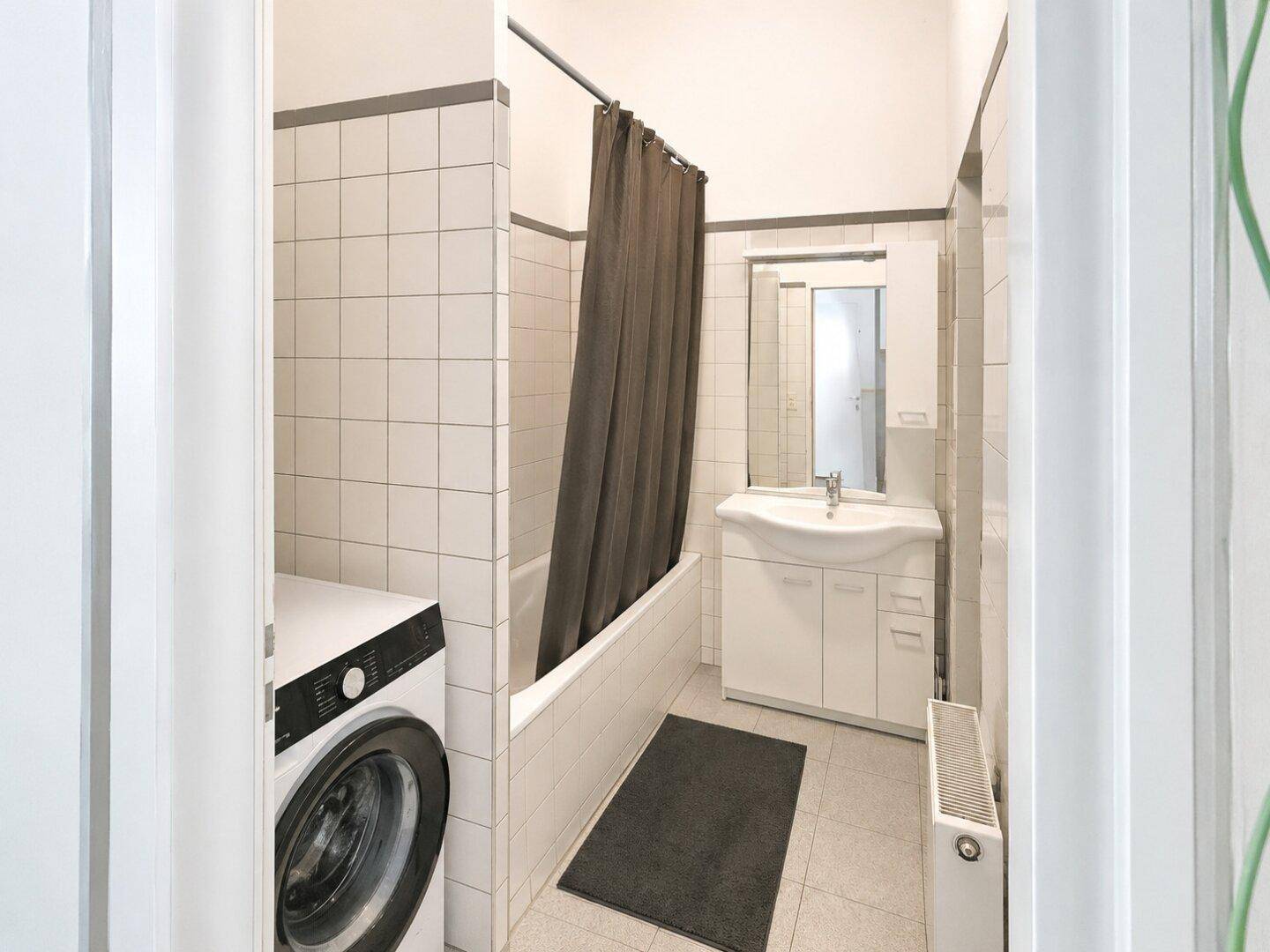 Helles Badezimmer mit Badewanne, Waschmaschine und weißen Wandfliesen.