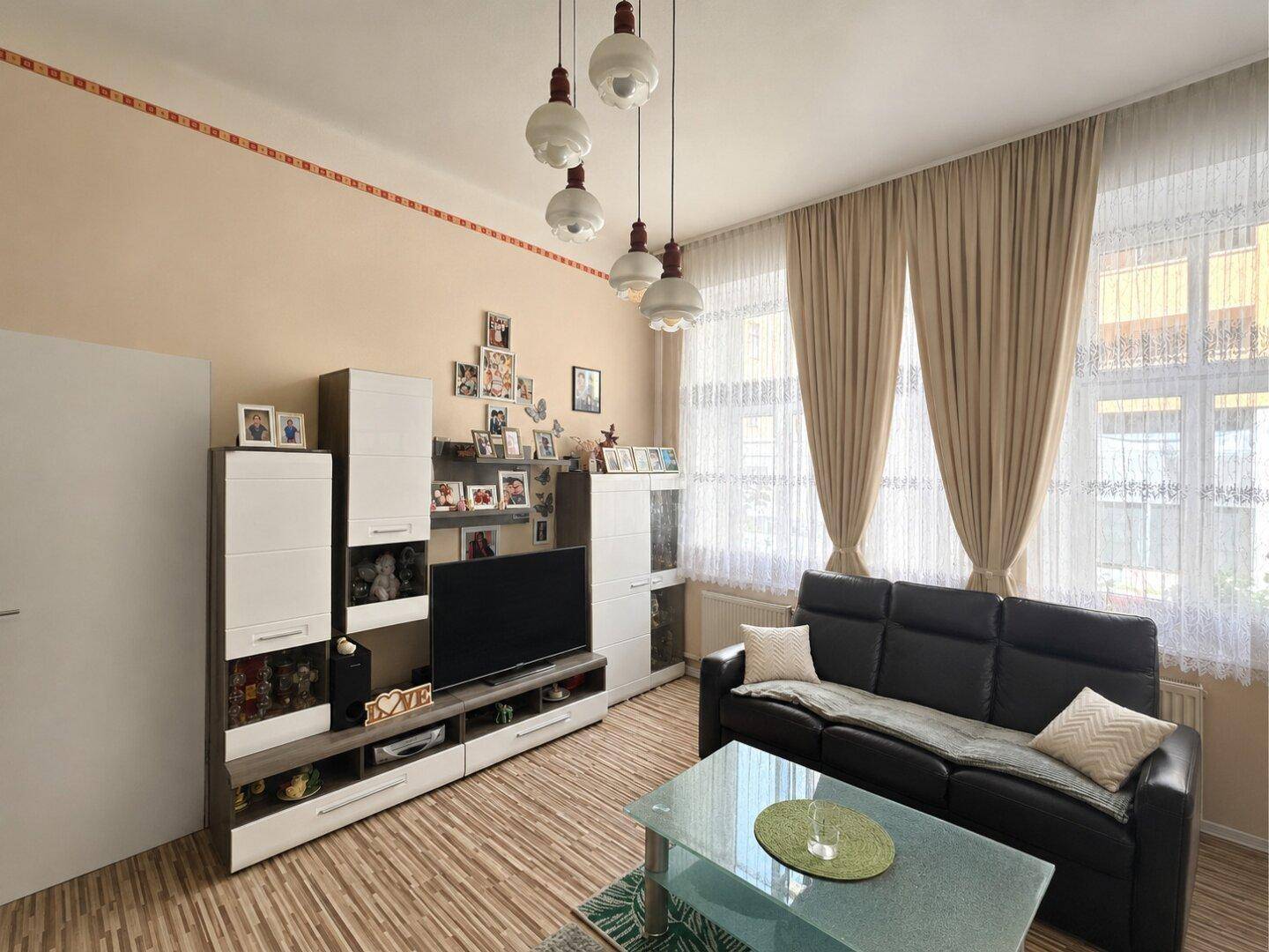 Helles Wohnzimmer mit modernem TV-Möbel, beigem Wandanstrich und großen Fenstern mit Vorhängen.