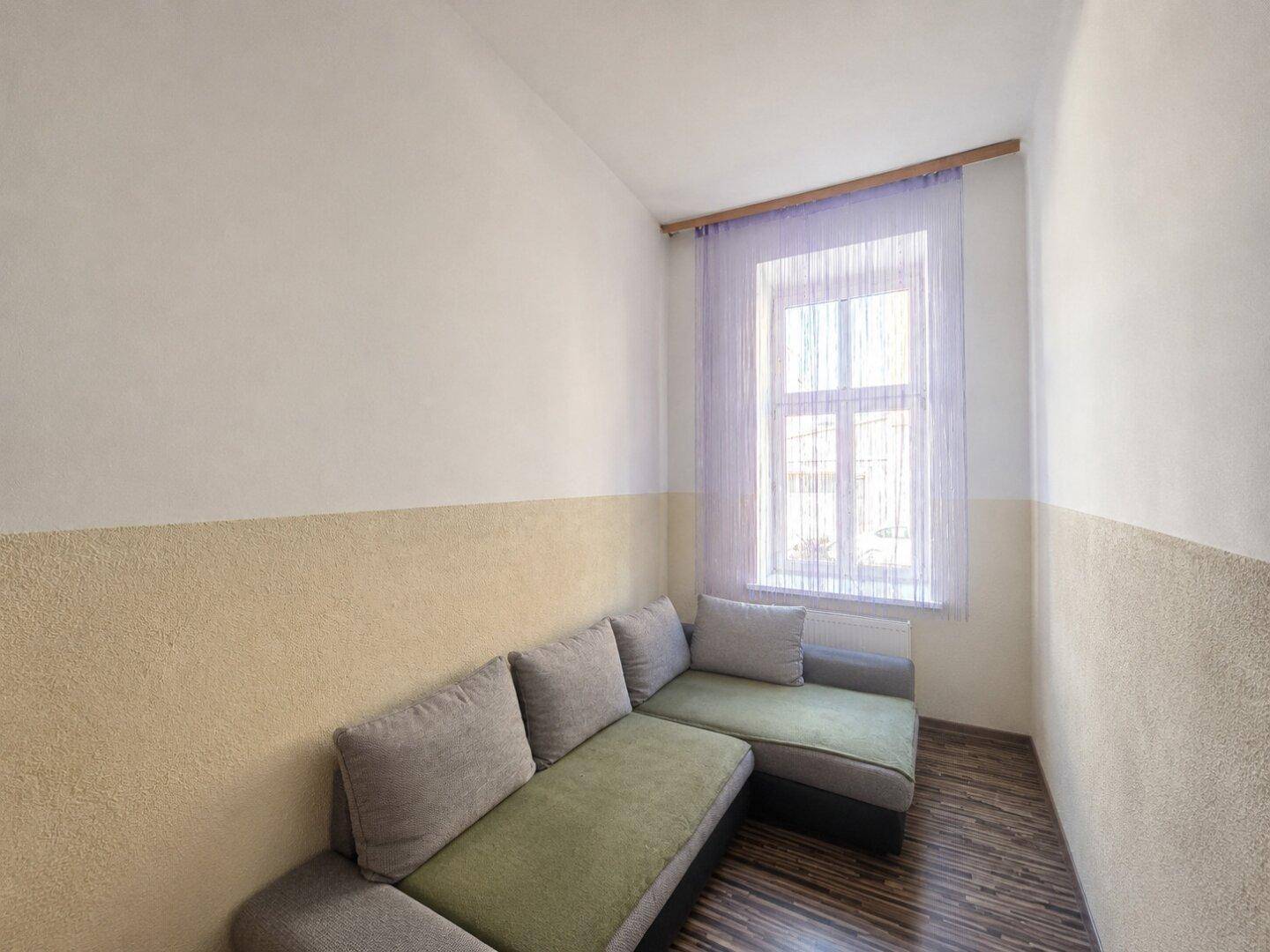 Kleines Zimmer mit Schlafsofa, zweifarbiger Wandgestaltung und einem Fenster mit lila Vorhang.