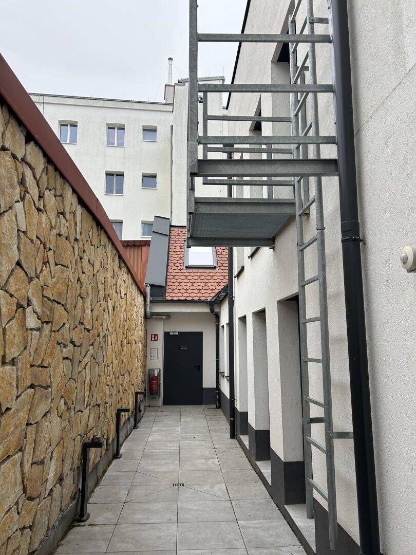 Schmaler Außenbereich zwischen Gebäuden mit Steinmauer und externer Metalltreppe.