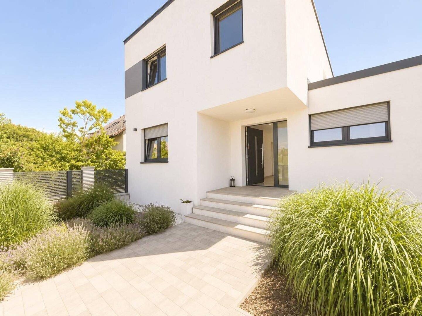 Moderne weiße Hausfassade mit dunklen Fenstern und Eingangsbereich, umgeben von gepflegter Bepflanzung.