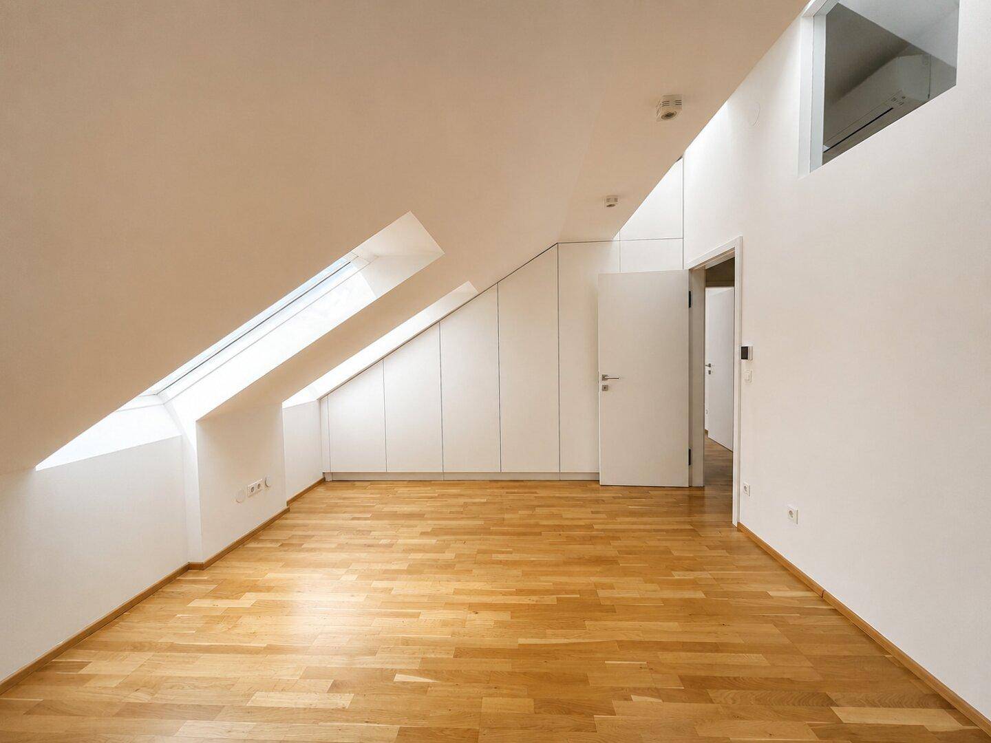 Heller Raum im Dachgeschoss mit schrägen Wänden, Dachfenstern und Holzboden.