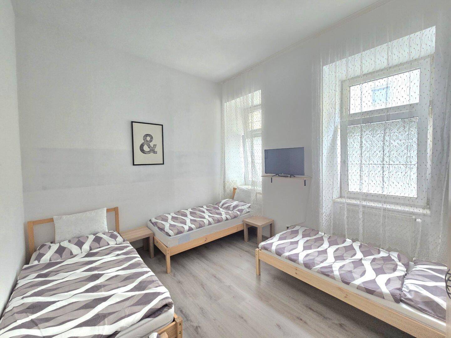 Helles Schlafzimmer mit drei Einzelbetten, kleinem Fernseher und zwei Fenstern mit Vorhängen.