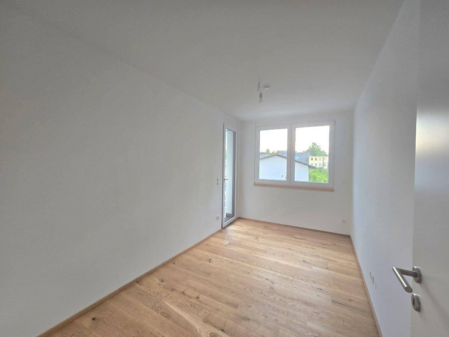 Leerer Raum mit Holzboden, weißen Wänden und Zugang zu einem Außenbereich mit Blick auf Dächer.