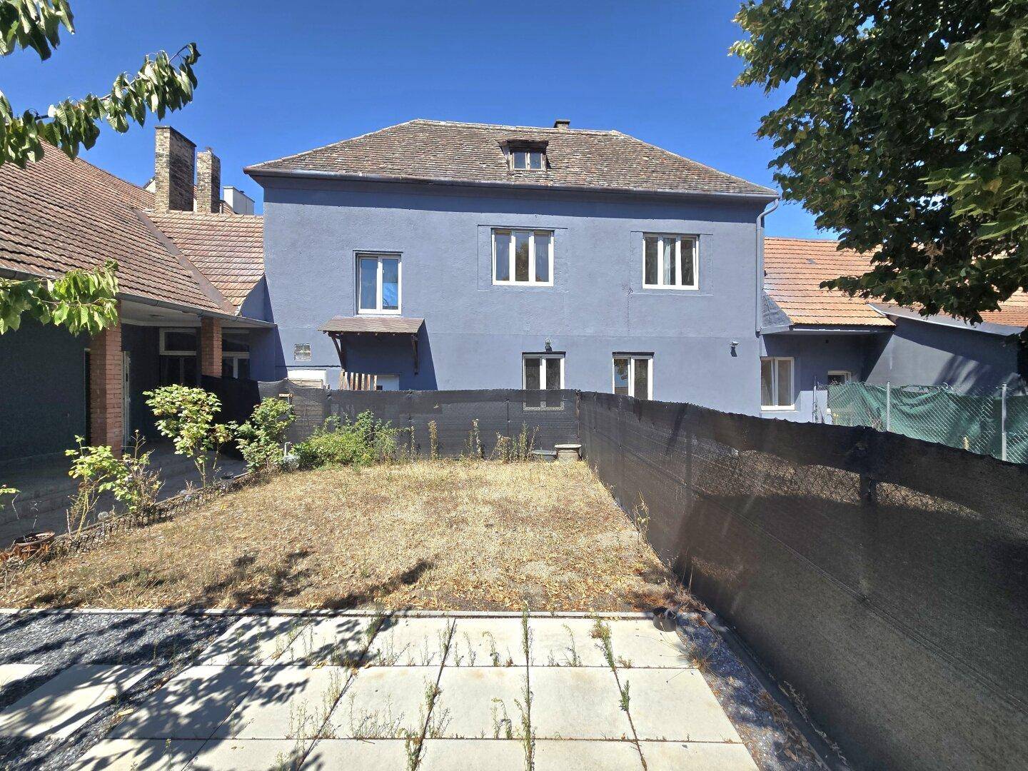 Rückansicht eines blaugrauen Gebäudes mit Gartenbereich und einer schwarzen Sichtschutzwand unter klarem Himmel.