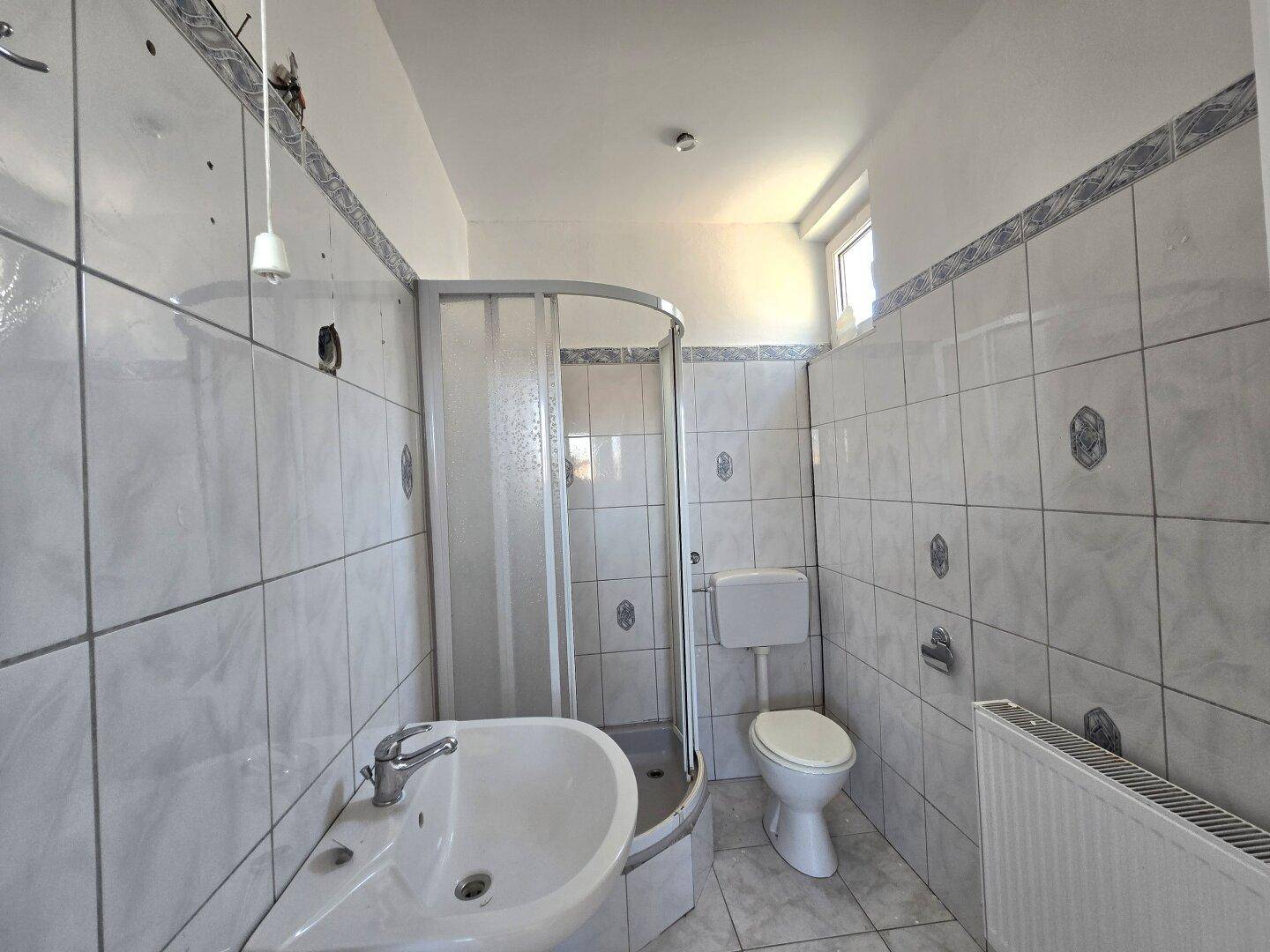 Gefliestes Badezimmer mit Duschkabine, WC, Waschbecken und einem Fenster für natürliche Belüftung.