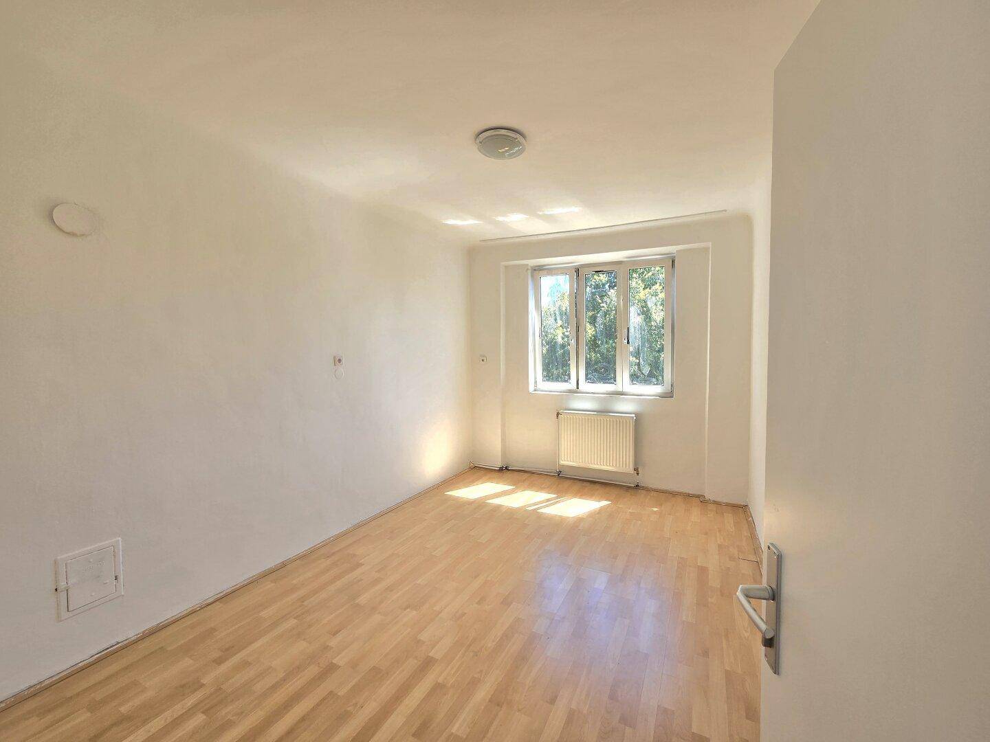 Helles Schlafzimmer mit Holzparkettboden und einem großen Fenster, das viel Sonnenlicht hereinlässt.