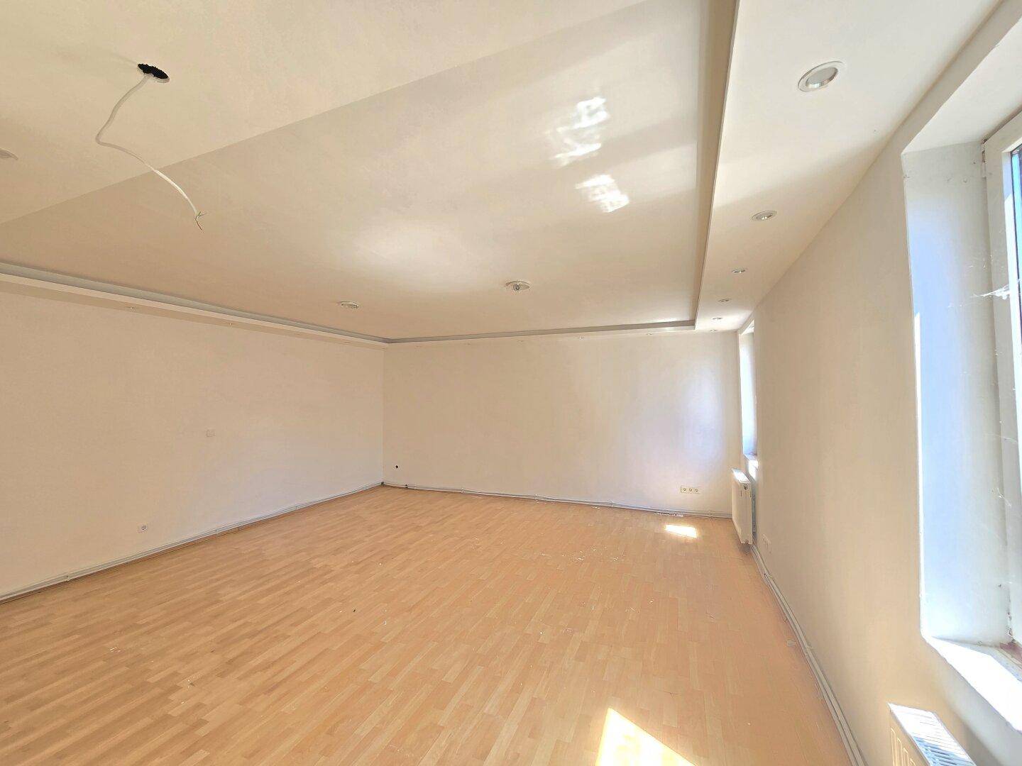 Geräumiger, heller Raum mit Holzparkettboden und einer abgehängten Decke mit integrierten Strahlern.