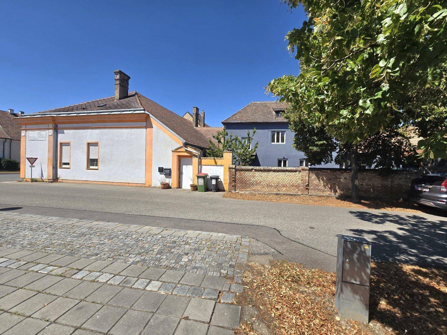 Ansicht des Gebäudes von der Straße mit einer Ziegelmauer und Bäumen im Vordergrund an einem sonnigen Tag.