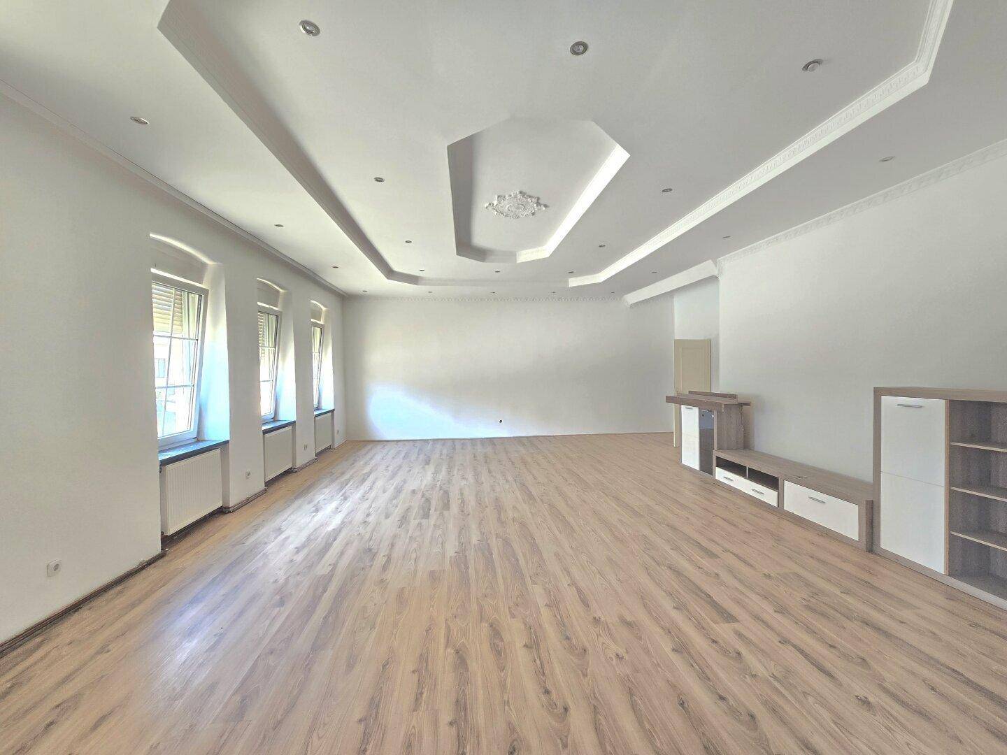 Geräumiges, helles Wohnzimmer mit modernem Laminatboden, dekorativer Deckenbeleuchtung und hohen Fenstern.