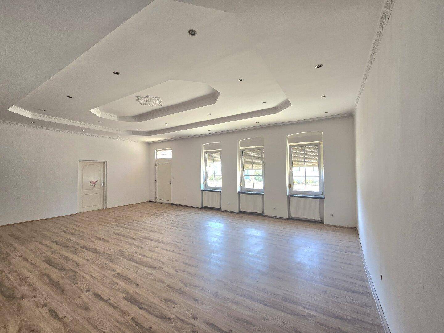 Großes, lichtdurchflutetes Wohnzimmer mit Laminatboden, eleganten Deckenleisten und mehreren Fenstern.