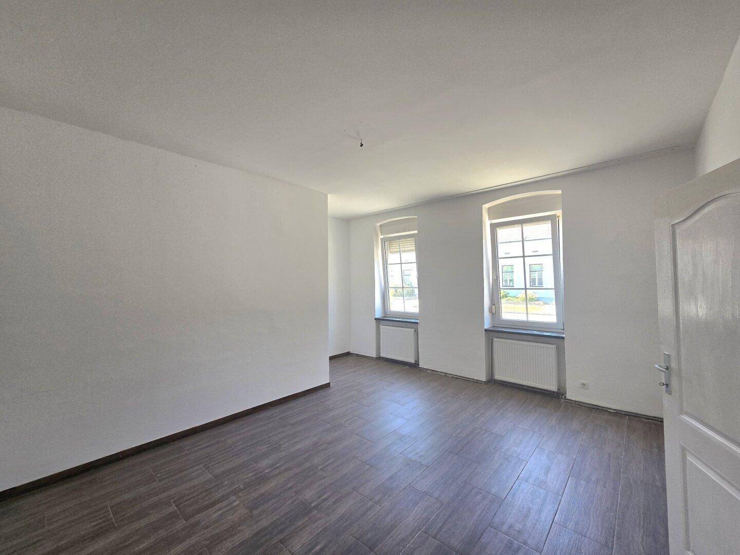 Helles Zimmer mit Laminatboden, zwei Fenstern und Heizkörpern unter den Fensterbänken, ideal als Schlafzimmer.