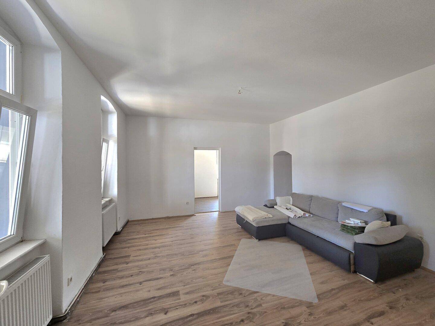 Gemütliches Wohnzimmer mit Laminatboden, einem Ecksofa und großen Fenstern, die viel Licht hereinlassen.