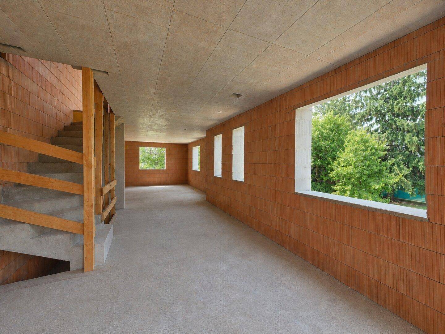 Unfertiger Innenraum eines Neubaus mit unverputzten Ziegelwänden, Betonboden und Treppe, bietet Blick ins Grüne.