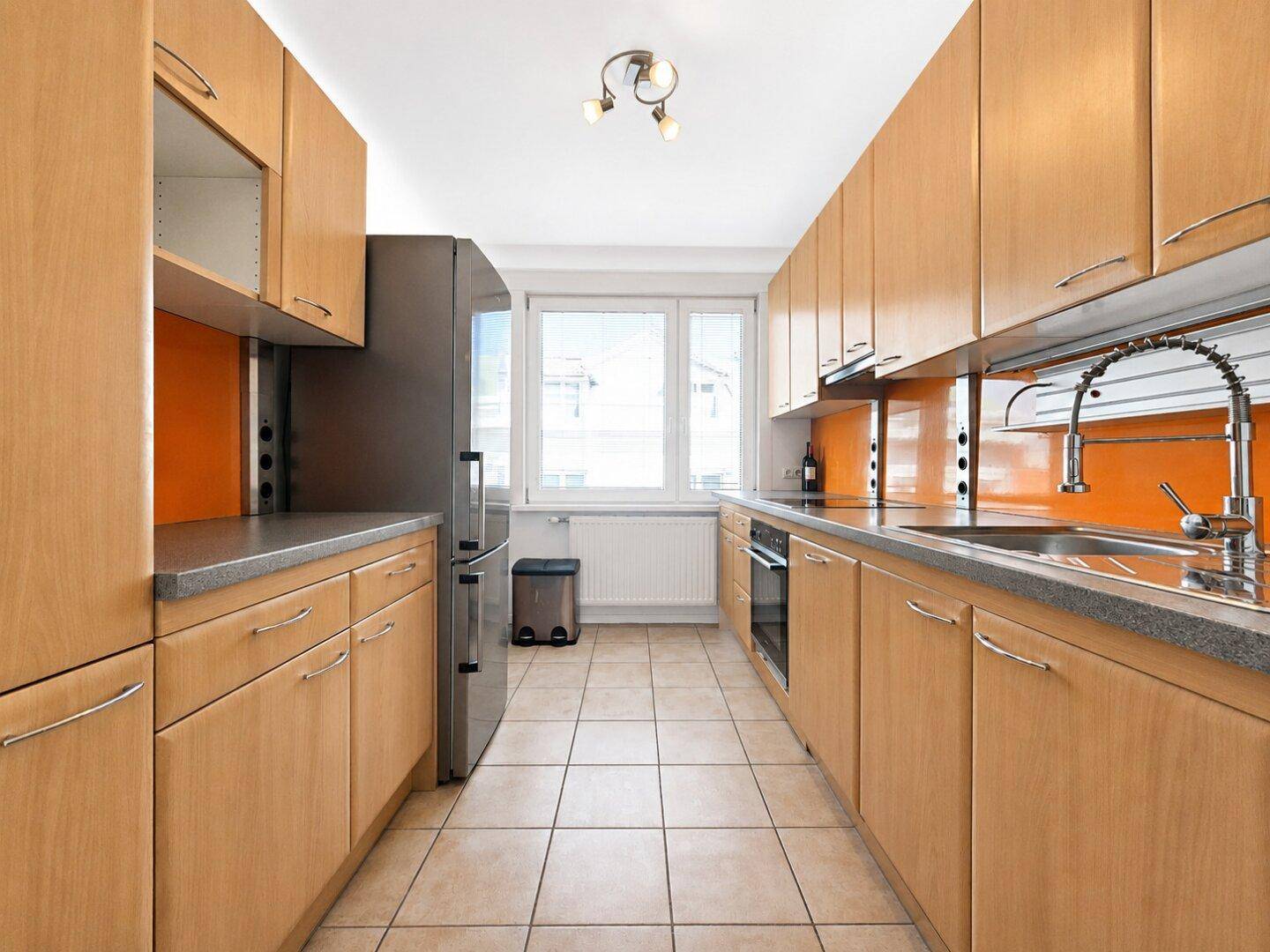 Funktionale Küche mit hellen Holzschränken, orangefarbenem Spritzschutz und Fliesenboden.
