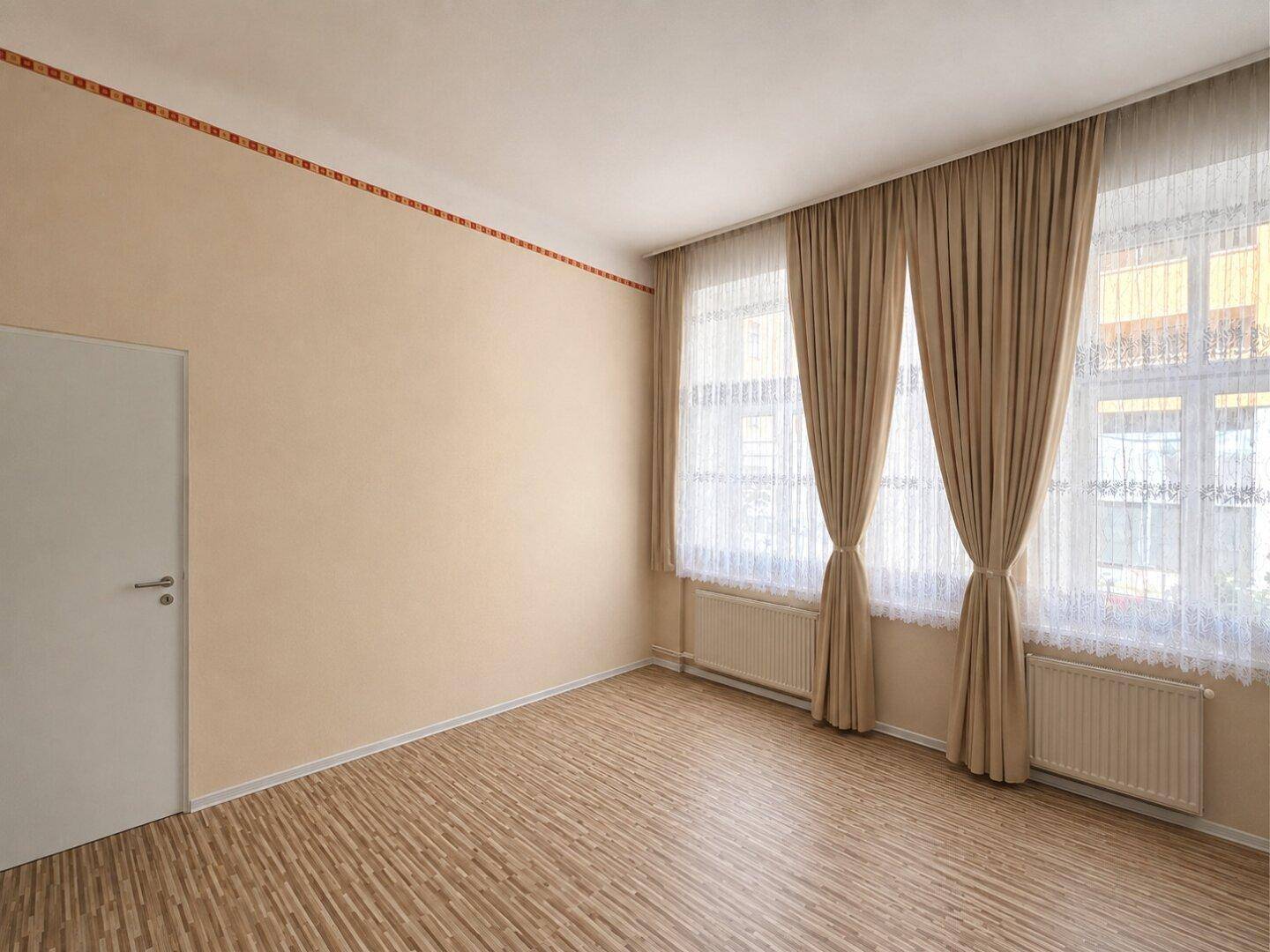 Heller Raum mit beigen Wänden, gemustertem Laminatboden und großen Fenstern mit Vorhängen.