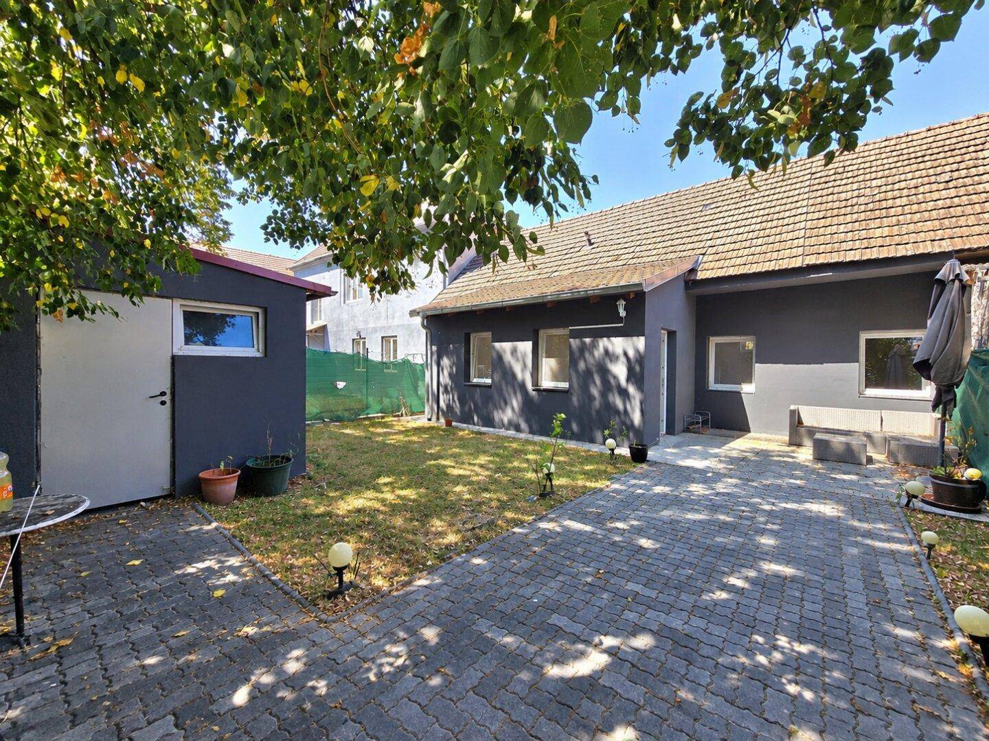 Hinterhof mit gepflastertem Weg, Rasenfläche, Gartenhaus und schattenspendendem Baum.