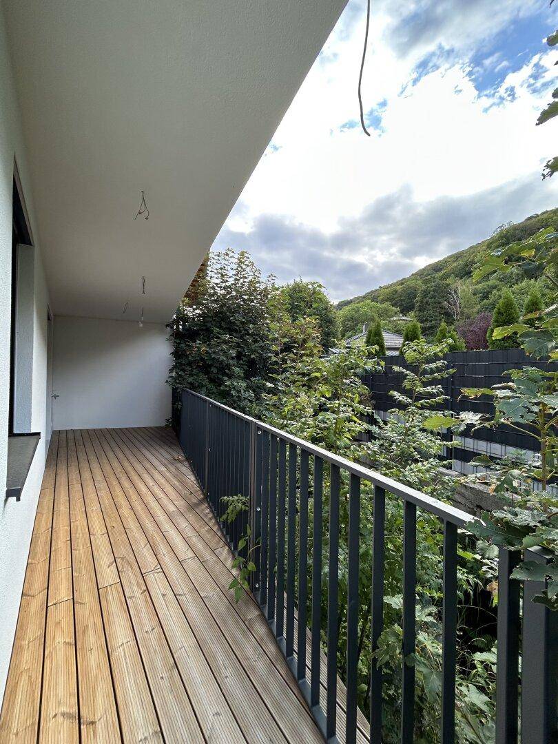 Langer Balkon mit Holzboden und Metallgeländer, umgeben von üppigem Grün und Bäumen.