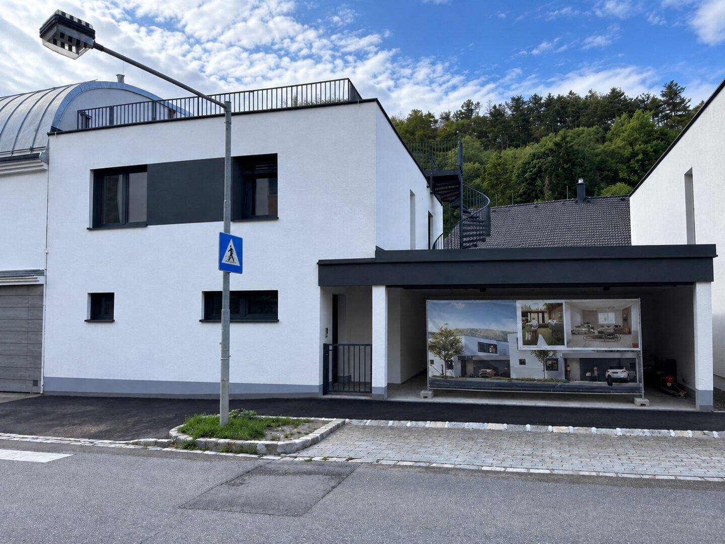 Moderne weiße Gebäudefassade mit dunklen Fenstern, Carport und Werbetafel für die Immobilie.