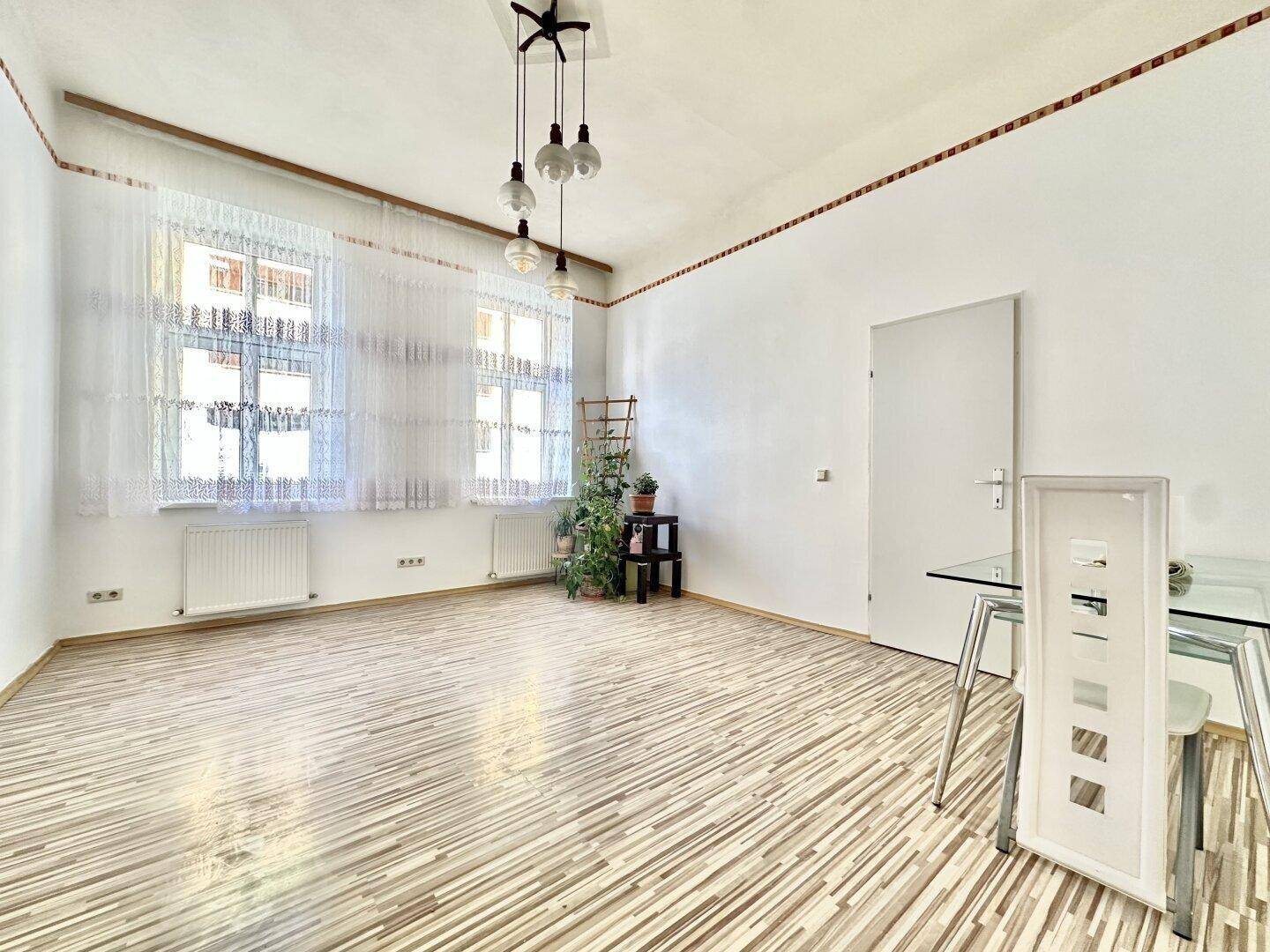 Heller Wohn-Essbereich mit großen Fenstern, hellem Laminatboden und moderner Deckenleuchte.