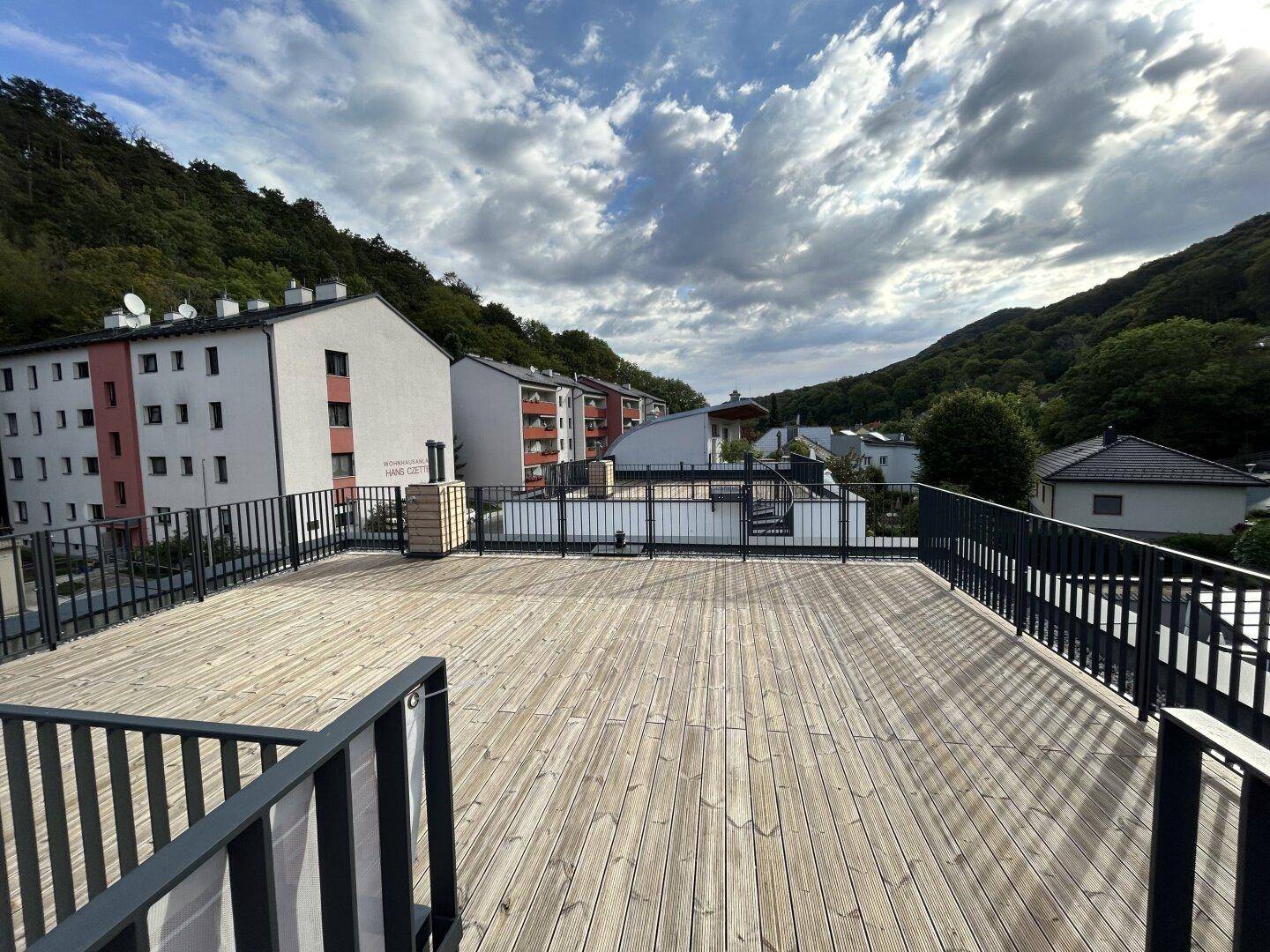 Weitläufige Dachterrasse mit Holzdielen und Blick auf Wohngebäude sowie bewaldete Hügel.