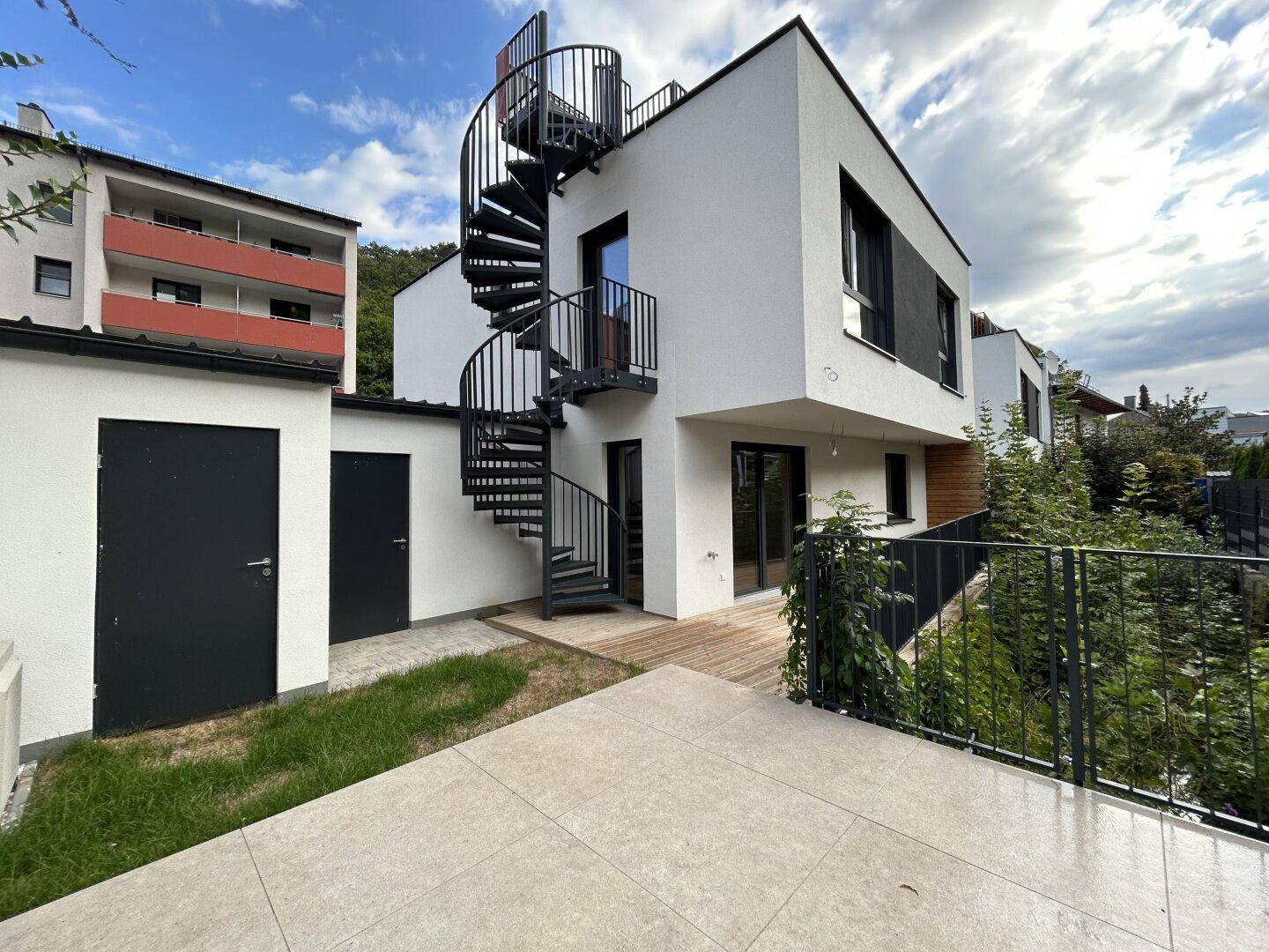 Seitliche Ansicht des modernen Gebäudes mit Außentreppe und kleiner gepflasterter Terrasse.