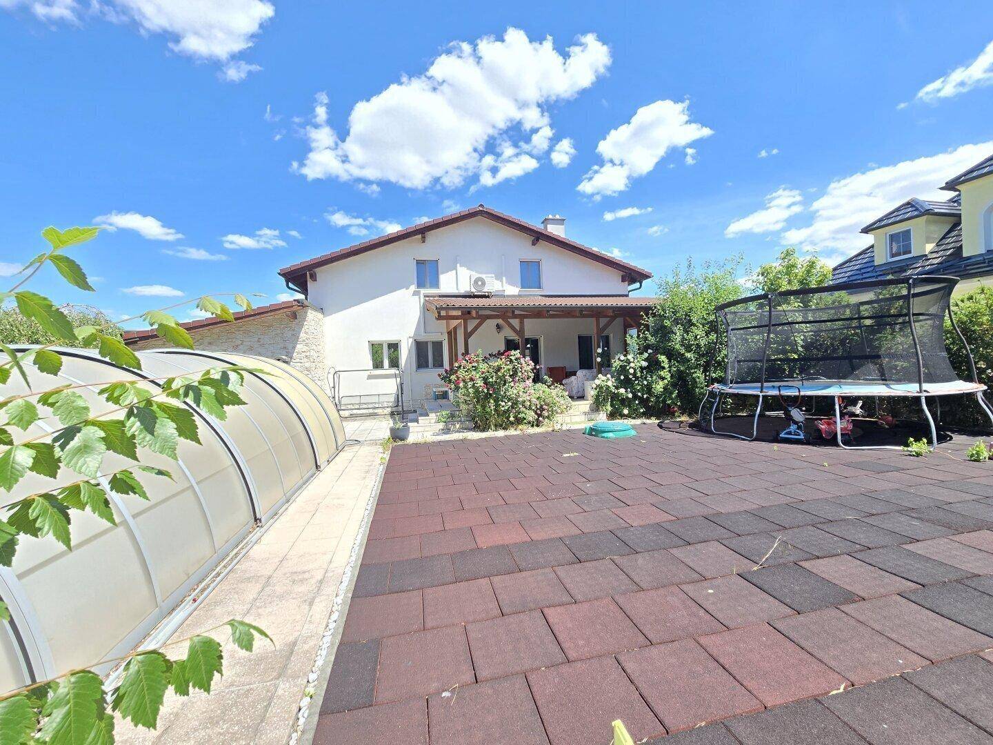 Haus mit großem Garten, überdachter Terrasse, Trampolin und Poolabdeckung unter blauem Himmel.