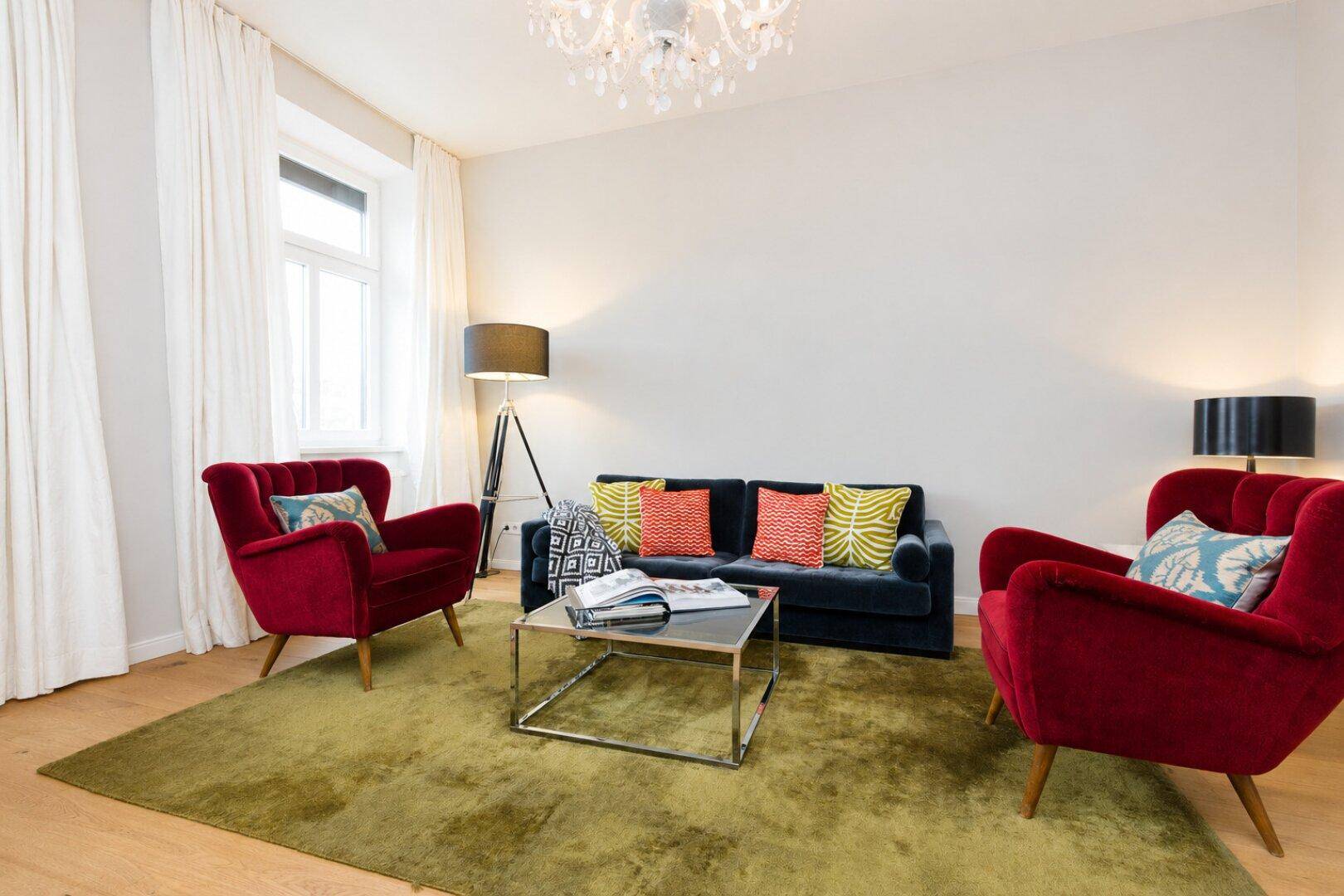 Geräumiges Wohnzimmer mit stilvollen roten Sesseln, einem blauen Sofa und einem auffälligen Kronleuchter.
