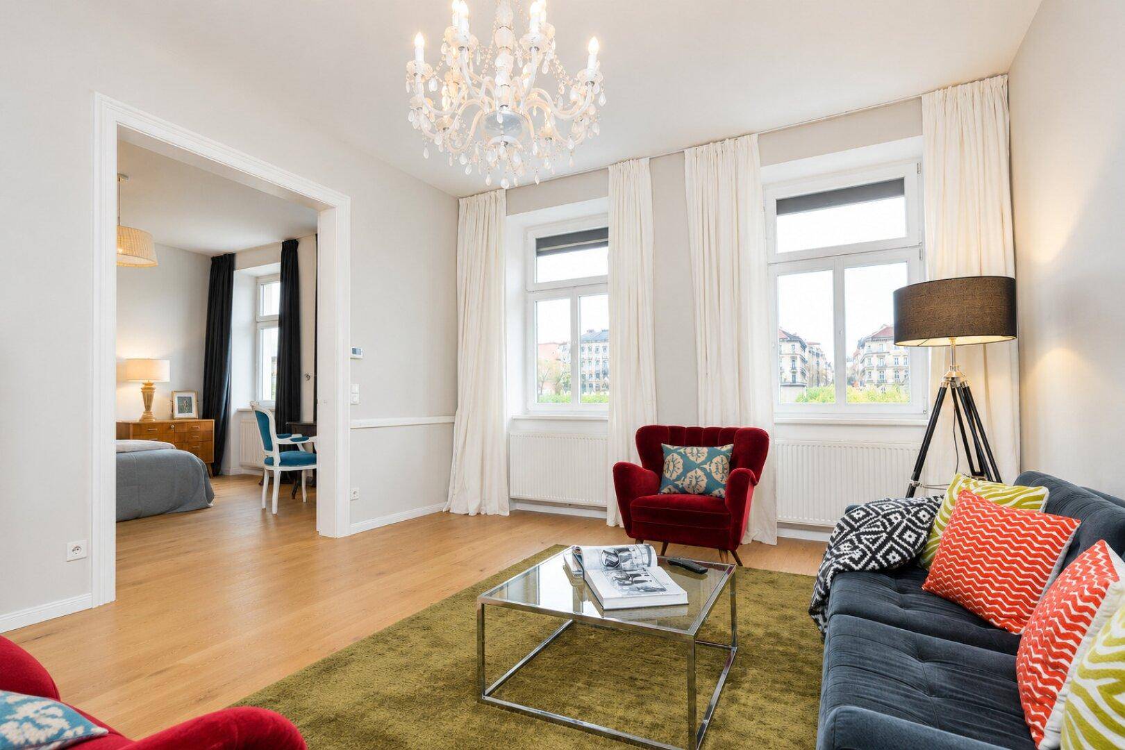 Helles Wohnzimmer mit bequemen Sitzmöbeln, Kronleuchter und Blick in ein angrenzendes Schlafzimmer.