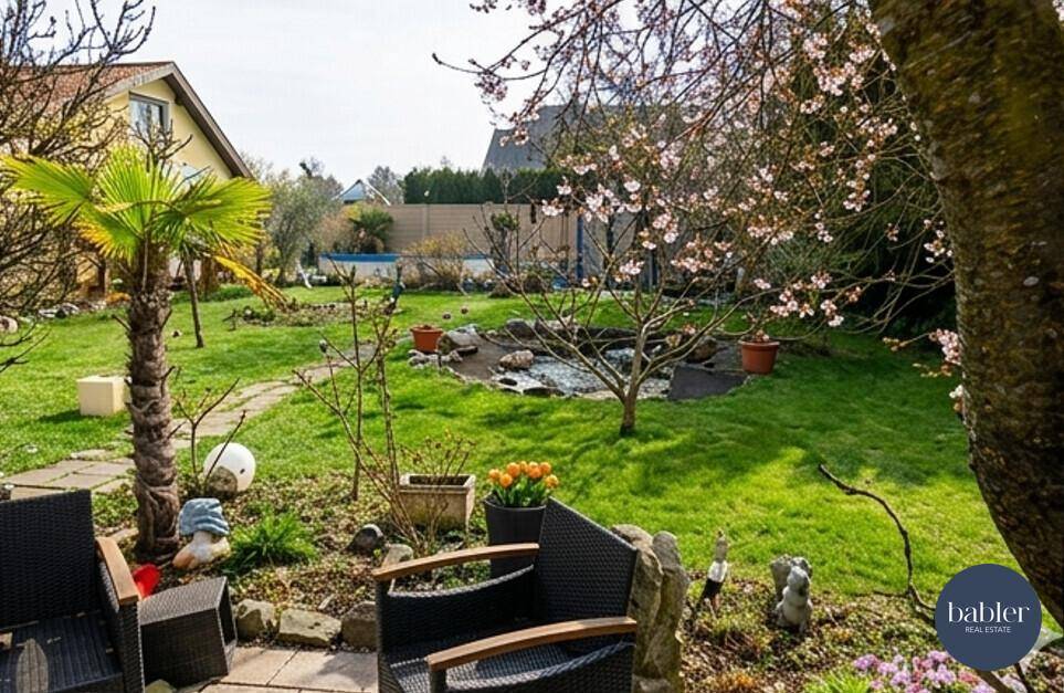 Großer Garten mit Sitzbereich, Teich und blühenden Bäumen unter blauem Himmel.
